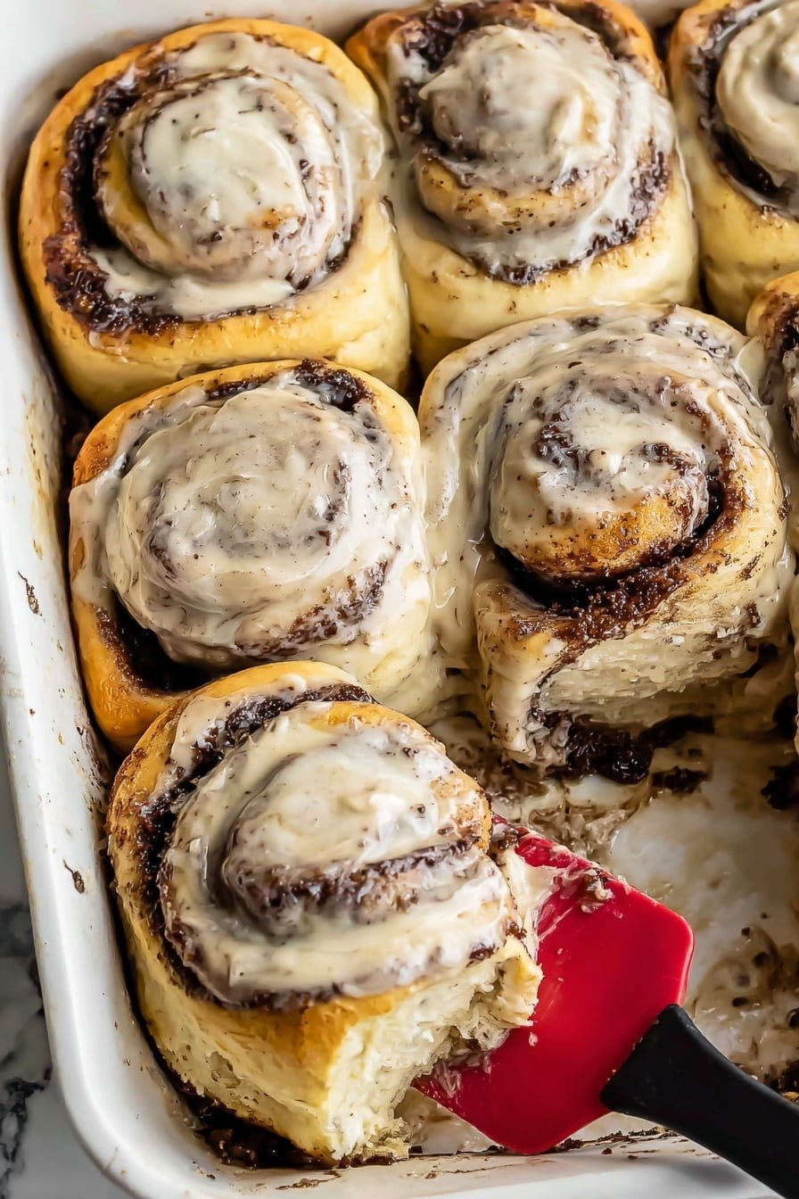 The image shows six large cinnamon rolls in a white baking dish, with golden-brown edges and thick swirls of dark cinnamon filling in between soft, fluffy layers of dough. Each roll is generously topped with a light beige cream cheese frosting that has tiny dark specks, spread smoothly over the spirals. One roll is being lifted on a red silicone spatula with a black handle from the bottom right corner, revealing gooey cinnamon filling beneath. The background features a white marbled surface. Photo taken with an iphone --ar 2:3 --v 7