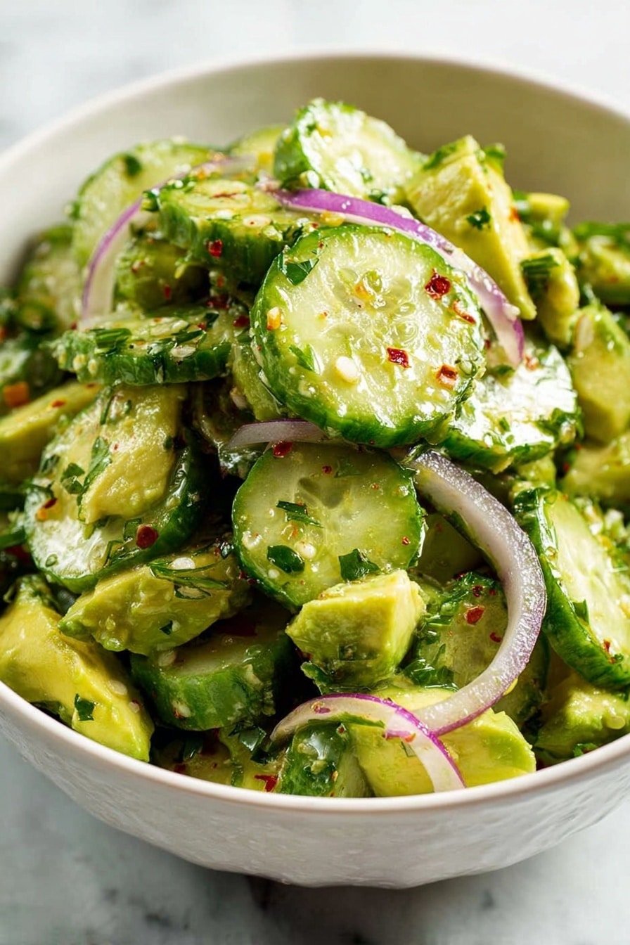 The image shows a white bowl filled with a fresh cucumber salad. The salad has two main layers: a base layer of green avocado chunks with a creamy texture, and a top layer of round cucumber slices that are green with pale centers and water droplets on the surface. Thin slices of purple onion are scattered throughout, adding a touch of color and texture. The salad is sprinkled with finely chopped green herbs and small pieces of red chili flakes, giving it a vibrant and fresh look. The white bowl sits on a white marbled surface. photo taken with an iphone --ar 2:3 --v 7