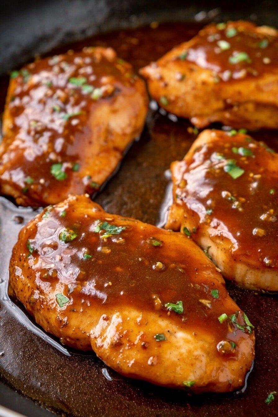The image shows four pieces of cooked meat, likely chicken, covered with a thick, shiny brown sauce. The sauce looks smooth with small bits and is spread unevenly over each piece, making them look moist and flavorful. On top of the sauce, small chopped green herbs are scattered, adding a touch of fresh color. The meat pieces are placed closely on a dark cooking surface with a slight shine from the sauce around them. The texture of the meat is tender and slightly uneven, showing it was cooked well. photo taken with an iphone --ar 2:3 --v 7