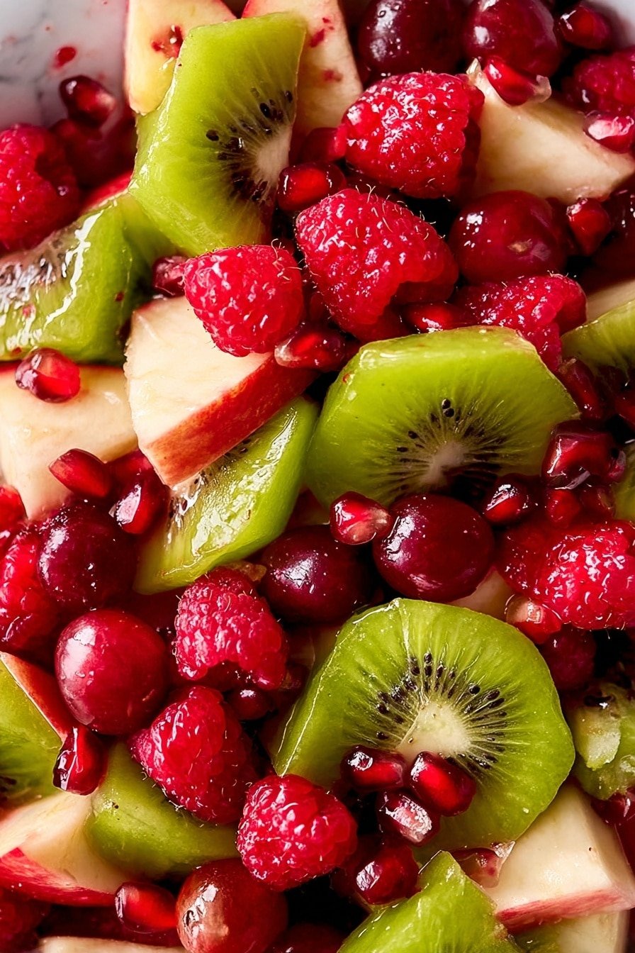 A white bowl filled with a colorful fruit salad containing at least five layers of mixed fruits. The bottom layer shows chopped red apple pieces with white flesh and bright red skin. On top of this layer are scattered glossy red grapes and green grapes. Above the grapes, there are thin green slices of kiwi fruit with visible small black seeds around the edges. Fresh bright red raspberries are spread evenly over the salad, adding a textured look. The salad is sprinkled with small, shiny, dark red pomegranate seeds all over. The bowl is placed on a white marbled surface with a small branch of red berries and green leaves visible in the corner. Photo taken with an iphone --ar 2:3 --v 7