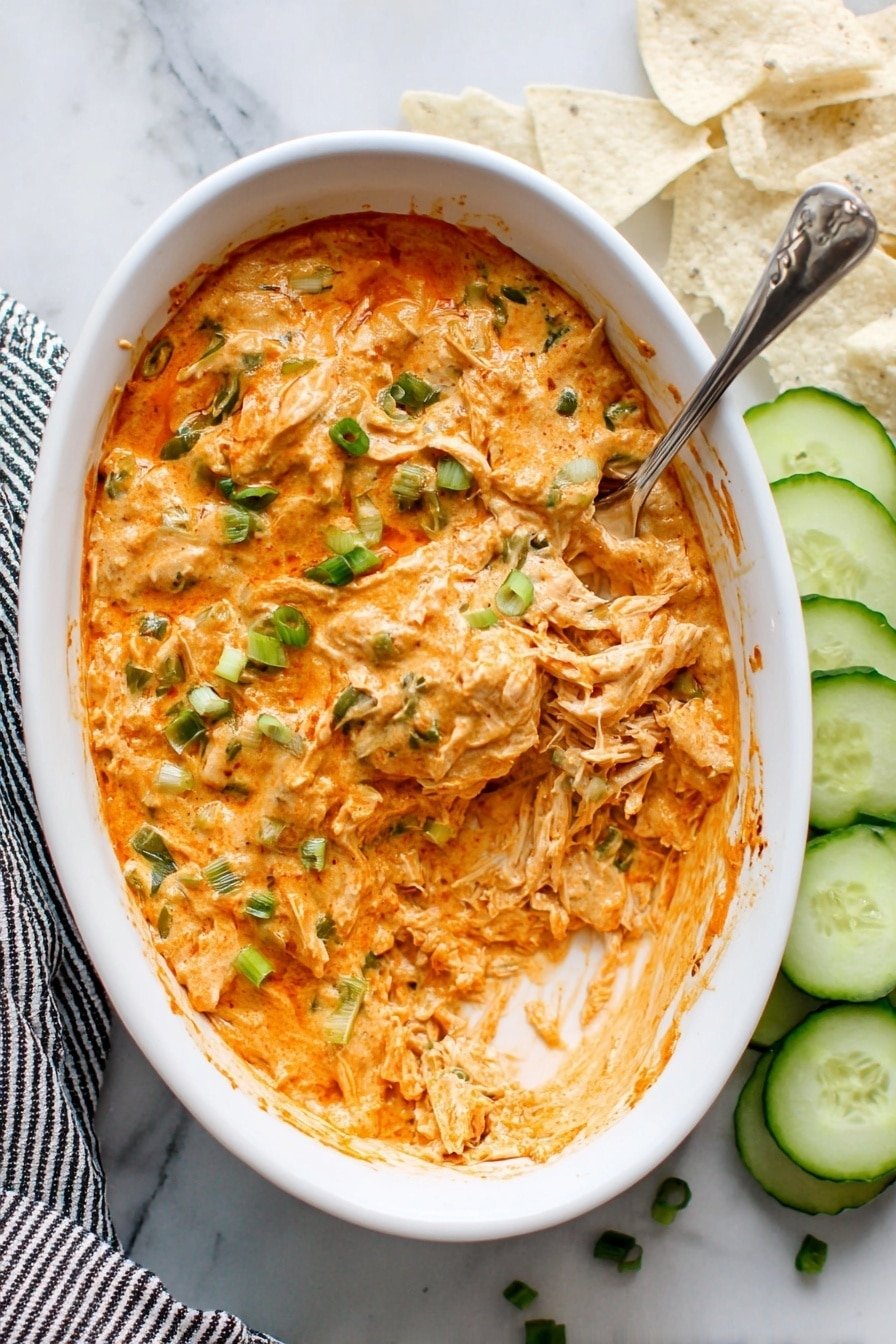 The image shows a white oval dish filled with a creamy, orange dip mixed with shredded pieces of cooked chicken or a similar protein, with visible green herbs and small bits of vegetables spread throughout. The top of the dip has a slightly browned, baked look with some green bits sprinkled on top, likely chopped scallions. A silver spoon is inside the dish, scooping up the dip, showing its thick and creamy texture with strands of the shredded ingredient. Surrounding the dish on a white marbled surface are neatly arranged cucumber slices and some white tortilla chips, with part of a black-and-white striped cloth napkin visible at the bottom left corner. photo taken with an iphone --ar 2:3 --v 7