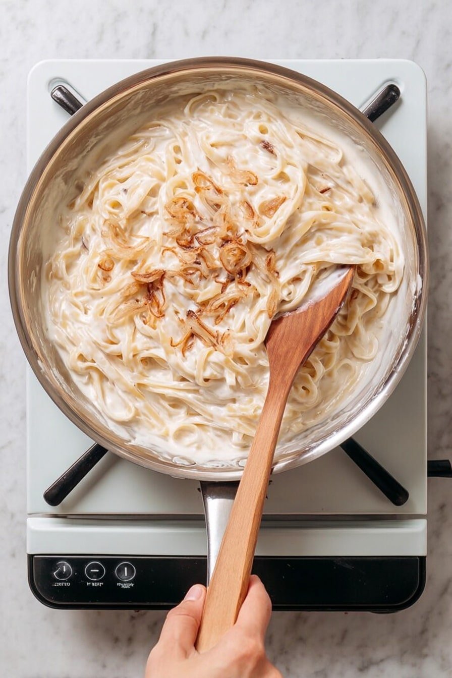 A large silver pan sits on a simple white electric stove with black burners. Inside the pan are thick, creamy noodles with a white sauce fully coating them, mixed with some brown bits that add contrast. A wooden spoon, held by a woman's hand, is stirring the noodles, showing creamy sauce sticking to it. The scene is set on a white marbled surface that softly contrasts with the pan and the stove. photo taken with an iphone --ar 2:3 --v 7