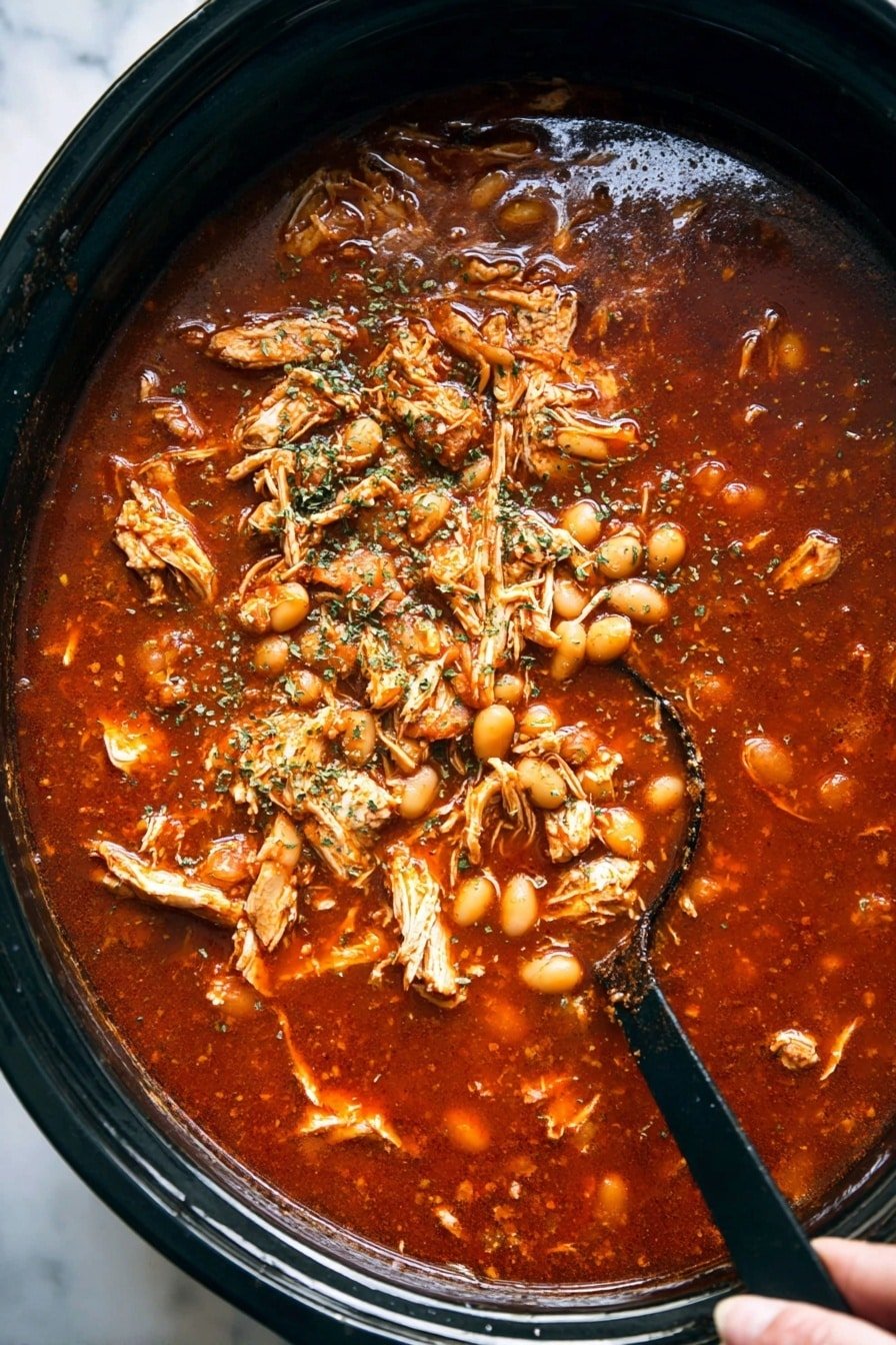 A large black pot filled with a thick red tomato-based stew containing shredded pieces of cooked chicken and small white hominy kernels scattered throughout. The stew is topped with dried green herbs sprinkled over the surface. A black ladle sits partially submerged on the right side of the pot, with a woman's hand holding it. The background surface is a white marbled texture photo taken with an iphone --ar 2:3 --v 7