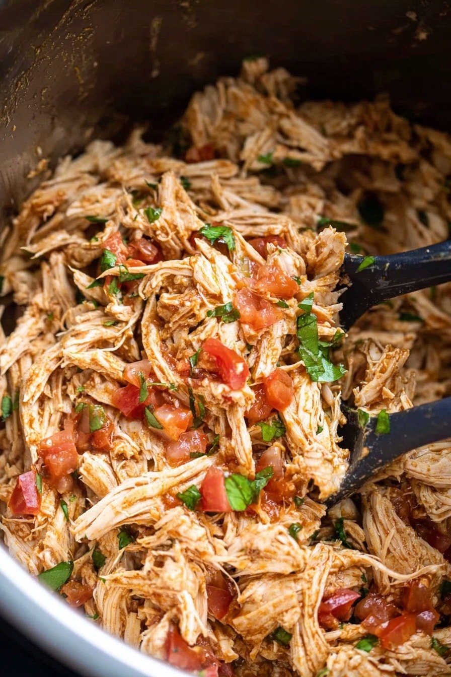 The image shows a close-up of a large stainless steel pot filled with shredded chicken mixed with small chunks of red tomato and green herbs scattered on top. The chicken looks tender with a light brown color mixed with the vibrant red of tomato pieces and bits of green herbs, giving a fresh appearance. Two black serving spoons are partially visible in the pot, creating slight texture contrast with their smooth surface. The shiny inside of the pot reflects light softly around the food. photo taken with an iphone --ar 2:3 --v 7