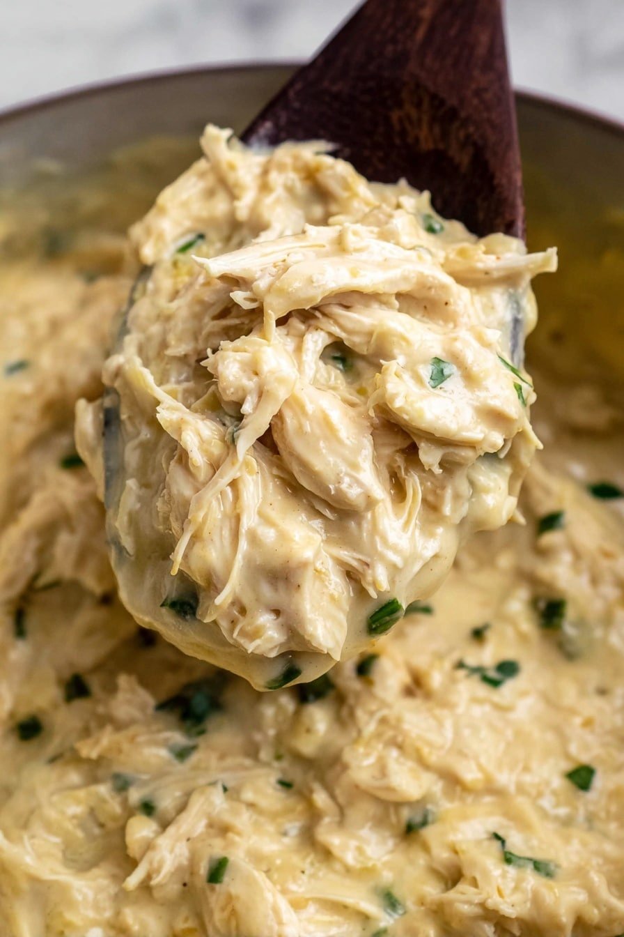 A close-up image shows a thick, creamy dish with a pale yellow color and a smooth, slightly chunky texture filled with shredded pieces of light beige chicken and tiny bits of green herbs. A dark wooden spoon lifts a scoop of the creamy mixture, prominently displaying the chicken strands and small green pieces. The dish appears hearty and dense, with a few scattered herb bits floating in the creamy base. The background shows more of the creamy mixture, all on a white marbled surface. Photo taken with an iphone --ar 2:3 --v 7