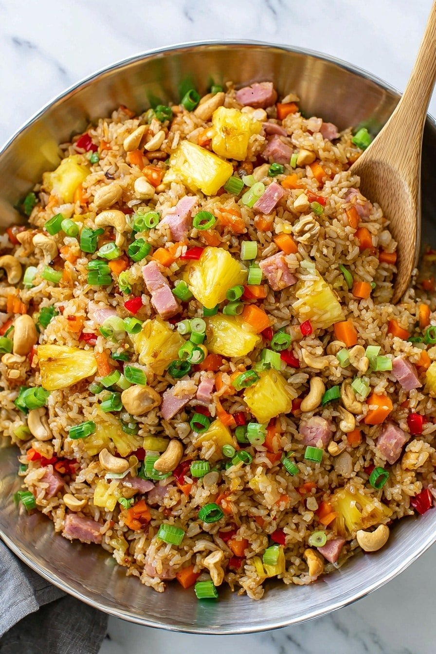 A large silver bowl filled with fried rice mixed with many colorful ingredients, including yellow pineapple chunks, small cubes of pink ham, bright orange carrot pieces, red bell pepper bits, green sliced scallions scattered on top, and whole cashew nuts. The rice grains are light brown, showing they are cooked with soy sauce. A silver spoon is partially buried in the rice on the right side of the bowl. The bowl sits on a white marbled surface. photo taken with an iphone --ar 2:3 --v 7