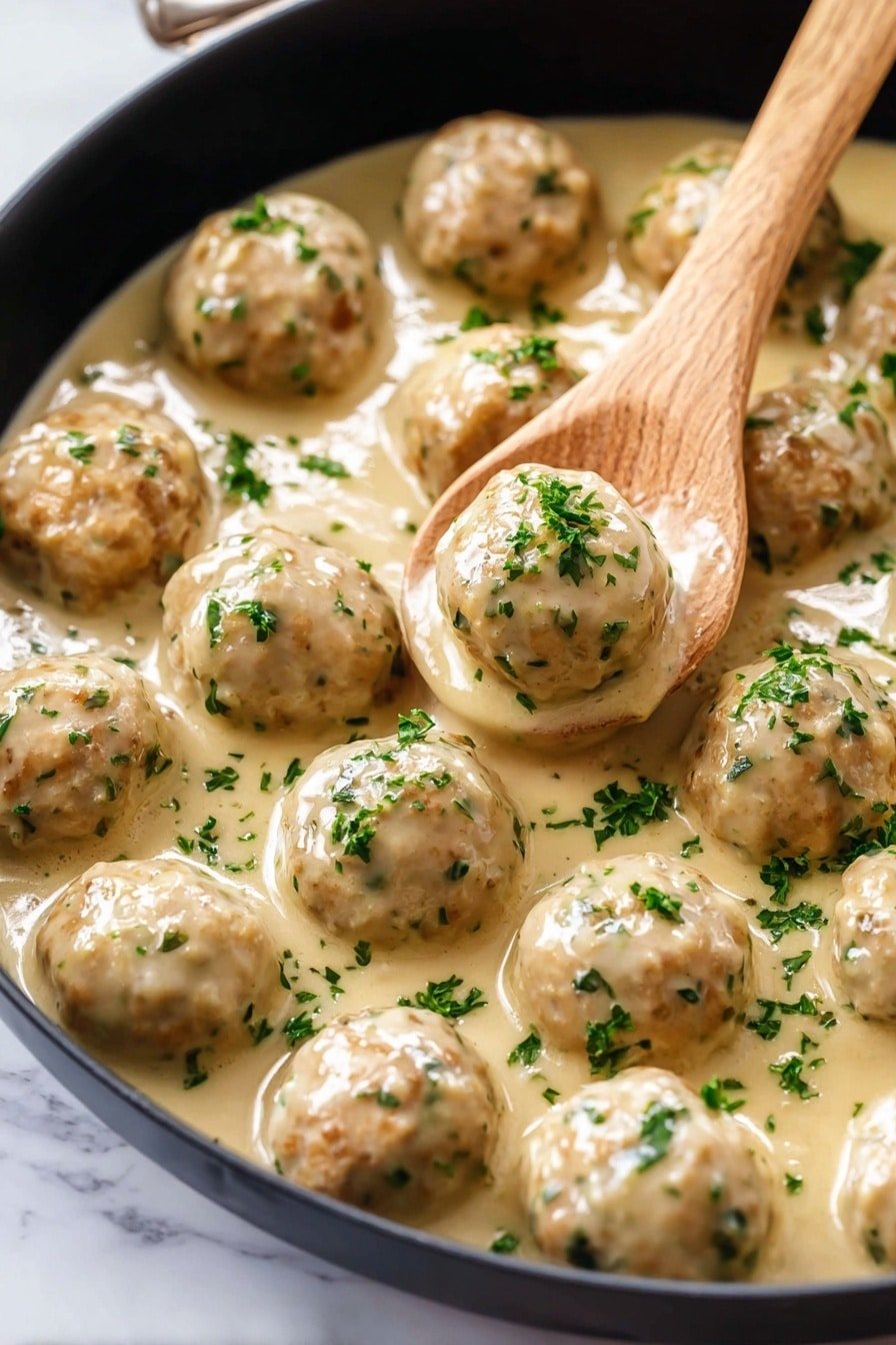 A close-up view of about twelve light brown meatballs with green herb bits mixed in, all evenly spread in a thick, creamy beige sauce inside a black pan. A wooden spoon is gently lifting one meatball near the center. The smooth sauce coats the meatballs, giving a shiny look, with small fresh chopped green herbs sprinkled inside and on top of everything. The pan sits on a white marbled surface. photo taken with an iphone --ar 2:3 --v 7