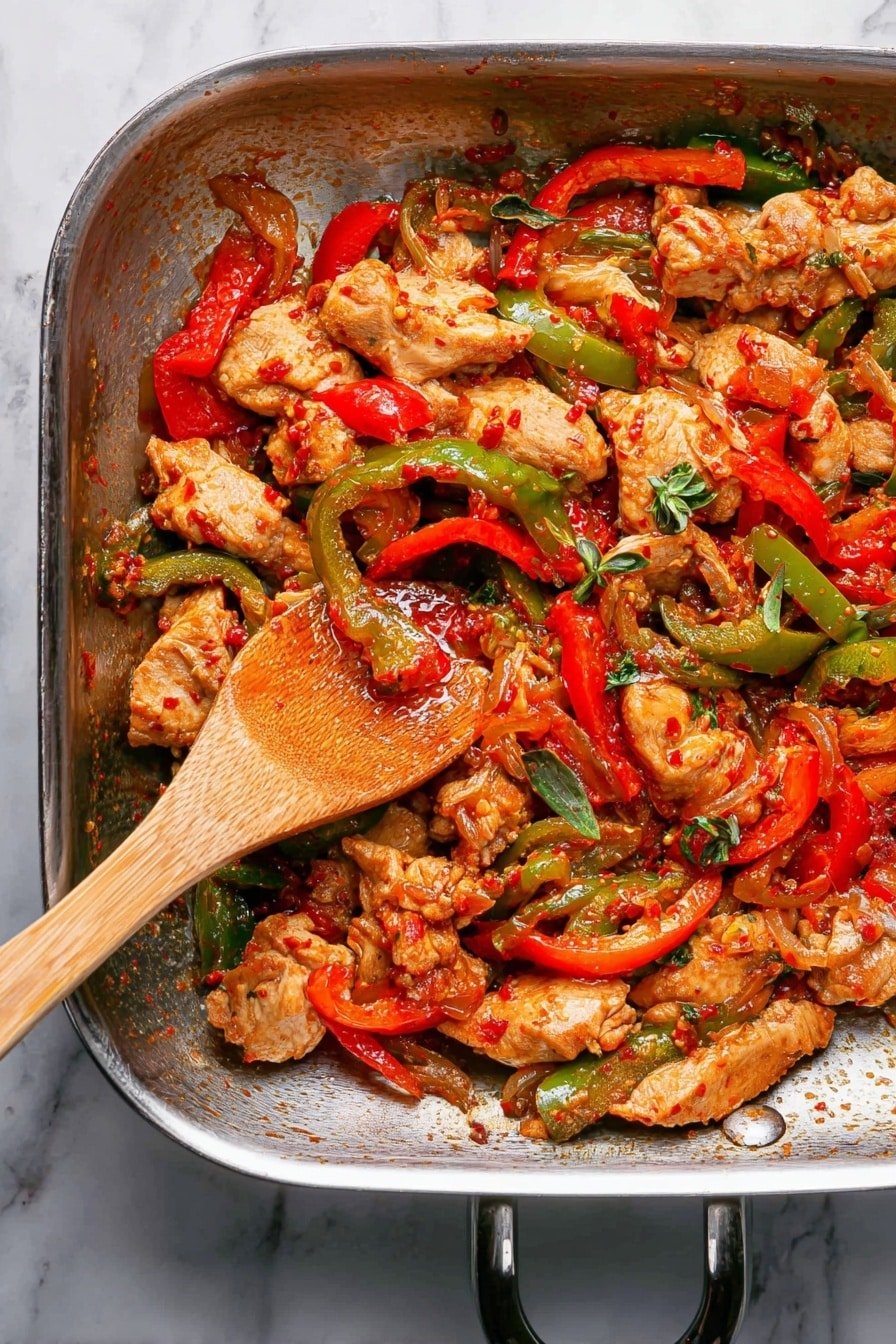 The image shows a metal pan filled with a cooked dish made of several layers: at the bottom, pieces of light brown cooked chicken with a slightly crispy look; mixed in are bright red and green sliced bell peppers adding bold colors; there are small bits of reddish sauce coating the ingredients, giving them a slightly shiny texture; some green herb leaves are scattered on top for a bit of freshness; a wooden spoon is resting in the pan, slightly stirring the food; the pan sits on a white marbled surface photo taken with an iphone --ar 2:3 --v 7