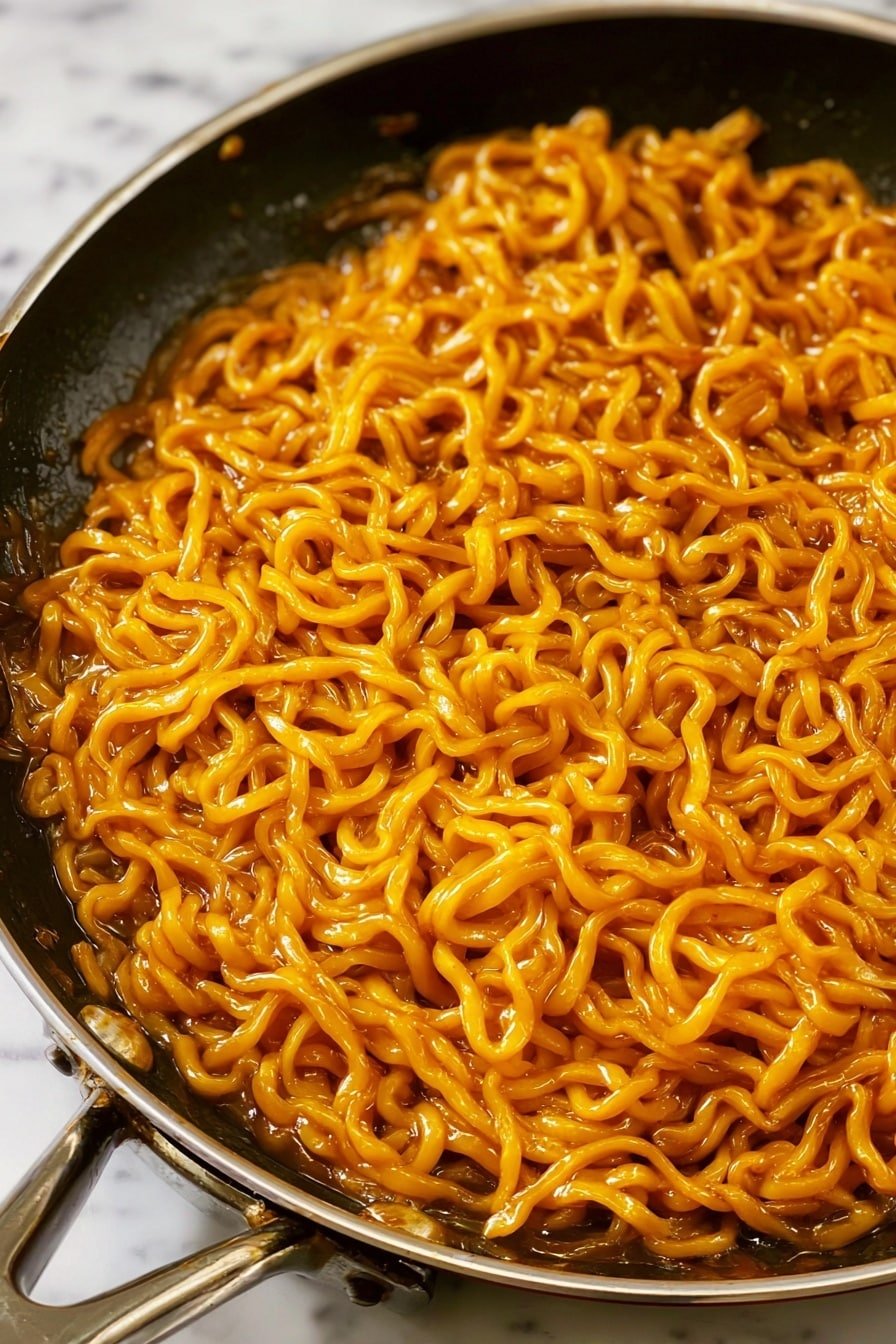 The image shows a close-up of a single layer of thick yellow noodles in a black pan with a silver handle. The noodles are well coated with a shiny, orange-brown sauce, giving them a moist and soft texture. The black pan contrasts with the bright color of the noodles, and the background is a white marbled texture. Photo taken with an iphone --ar 2:3 --v 7
