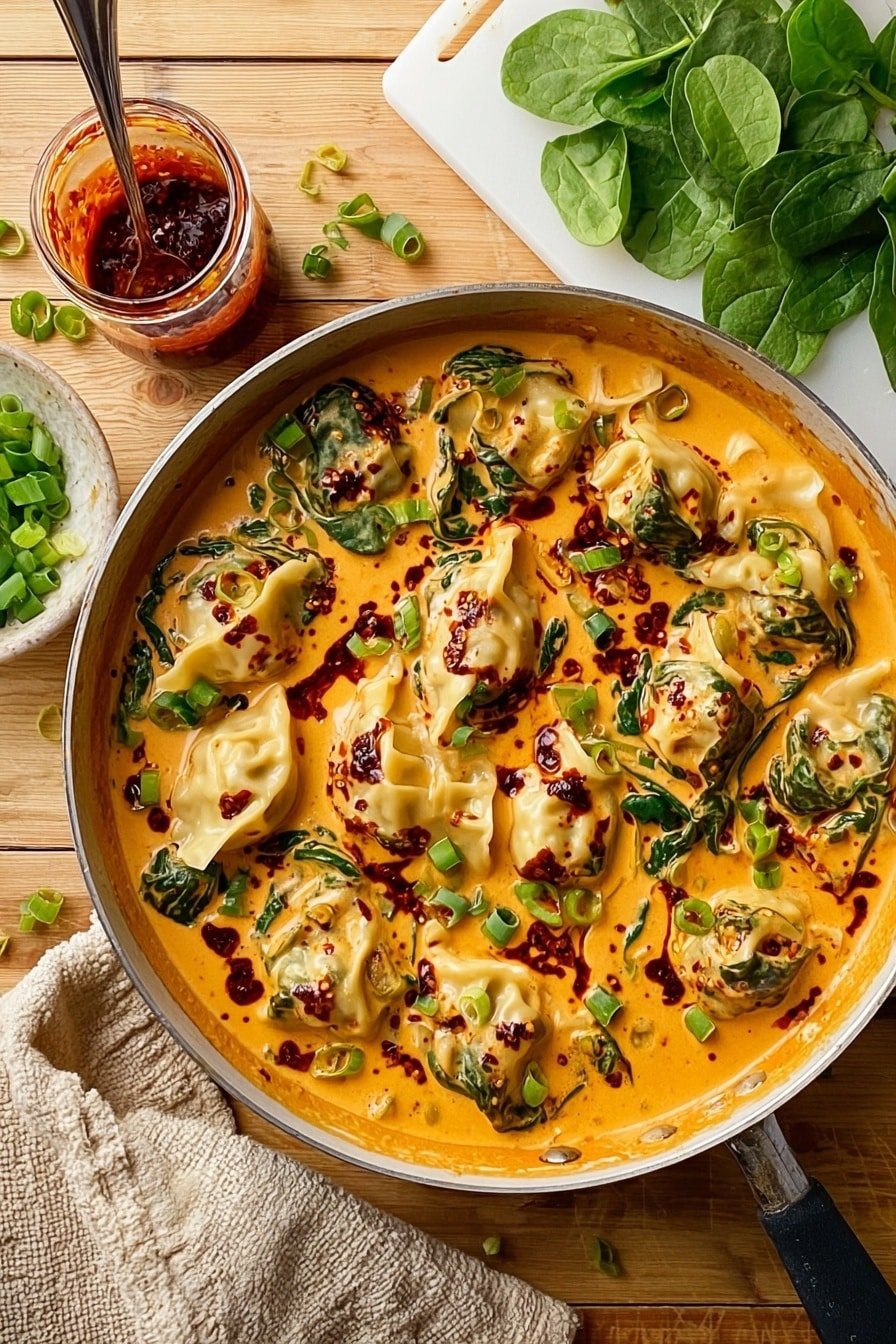 The image shows a round pan filled with about ten dumplings in a rich, creamy orange sauce. The sauce is smooth and thick, with dark red chili oil spots scattered across the surface. Fresh green spinach leaves are mixed inside the sauce, visible around and under the dumplings. Chopped green onions are sprinkled on top, adding a fresh bright green color. The pan rests on a wooden surface, with fresh spinach leaves placed on one side and a white cutting board with more chopped green onions on the other. A jar of chili sauce with a spoon inside is beside the pan, and a beige oven mitt is nearby. Photo taken with an iphone --ar 2:3 --v 7