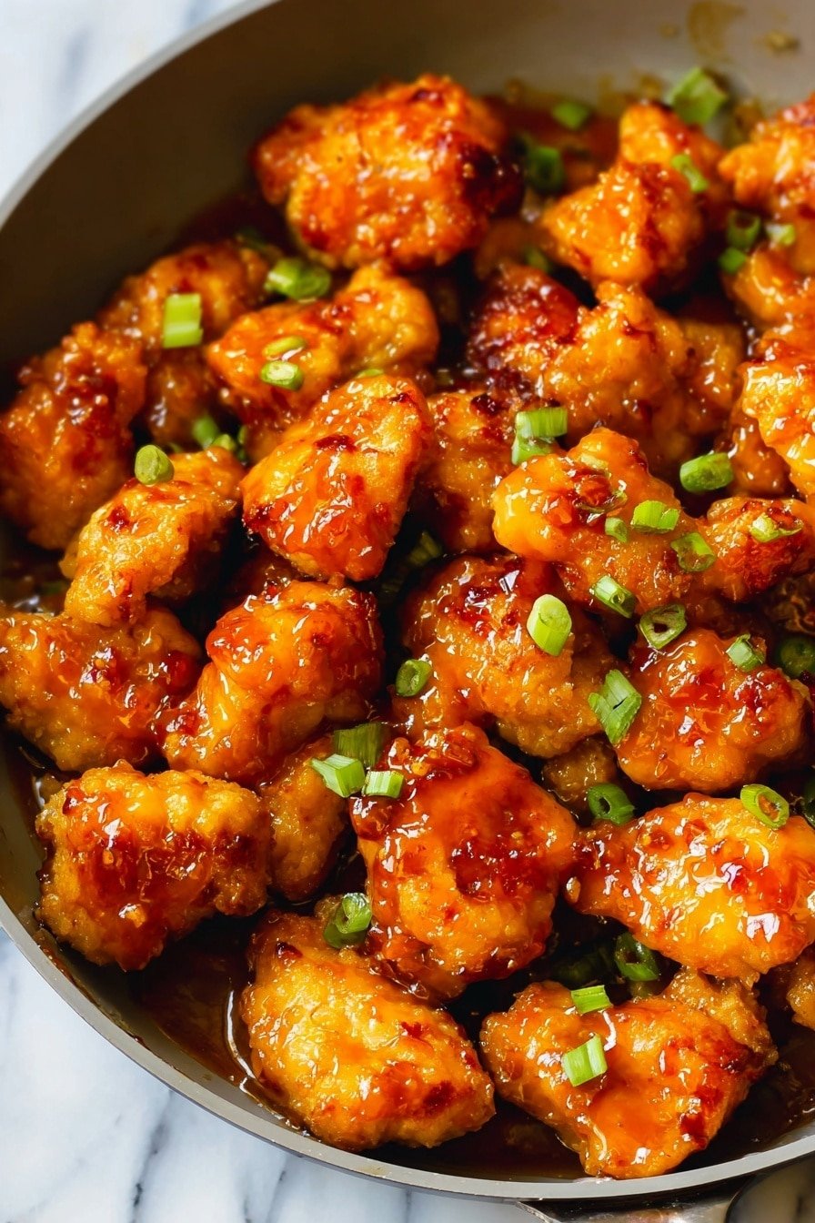 A pan filled with one layer of golden fried chicken pieces, each piece covered in a shiny orange glaze that glistens under the light. Small bright green chopped scallions are sprinkled evenly over the chicken, adding spots of fresh color. The chicken looks slightly crispy with some caramelized edges showing through the glossy sauce. The pan is round and white, resting on a white marbled surface. photo taken with an iphone --ar 2:3 --v 7