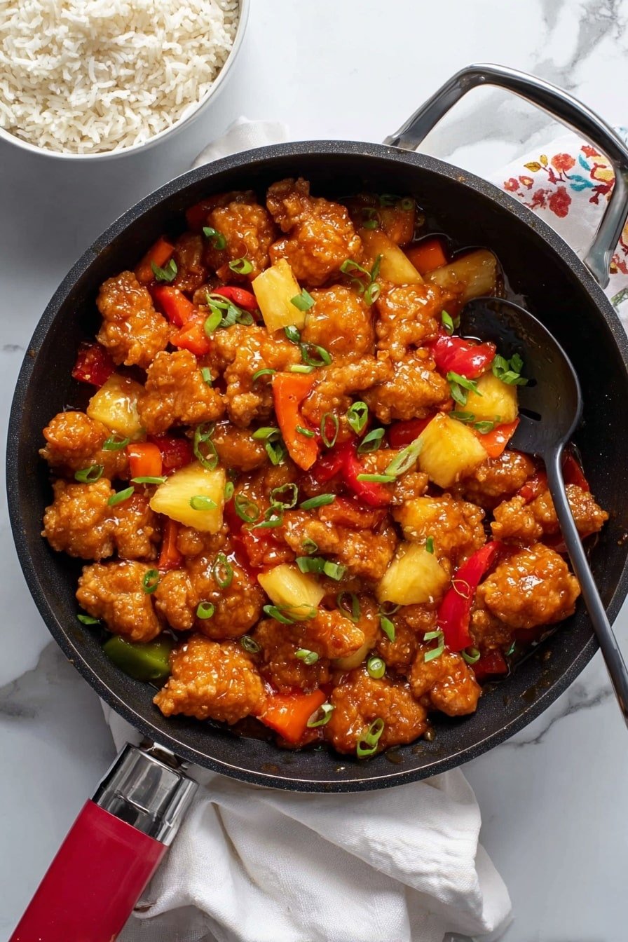 The image shows a large black pan filled with pieces of golden-brown fried chicken mixed with bright chunks of yellow pineapple and red and orange bell peppers. The chicken pieces are thick, coated in a shiny, sticky orange sauce that glistens under the light. Green chopped spring onions are sprinkled over the top, adding a pop of color. The pan rests on a white marbled surface with a white cloth napkin beside it. In the top left corner, there is a white bowl filled with plain cooked rice. The handle of the black pan is visible, with a red grip, and a black spoon lies inside the pan. Photo taken with an iphone --ar 2:3 --v 7