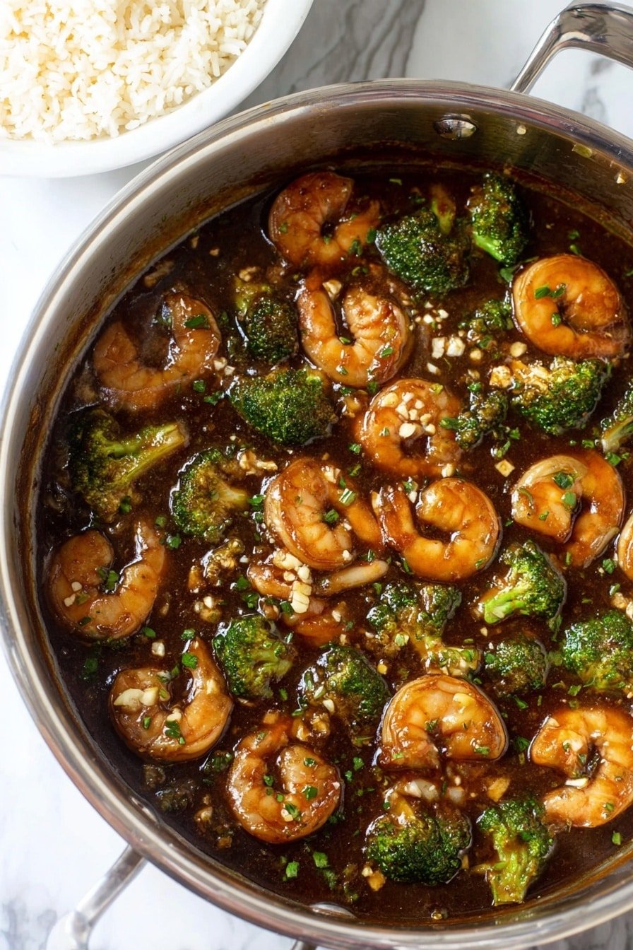 A close-up view of a silver pot filled with a thick, dark brown sauce covering medium-sized shrimp that are light brown with a glossy texture, and bright green broccoli florets scattered throughout. Small bits of chopped garlic and green herbs float on the surface of the sauce, which has a shiny finish. The pot is positioned on a white marbled surface, with a white bowl partly shown in the upper left corner filled with fluffy, white rice grains. photo taken with an iphone --ar 2:3 --v 7