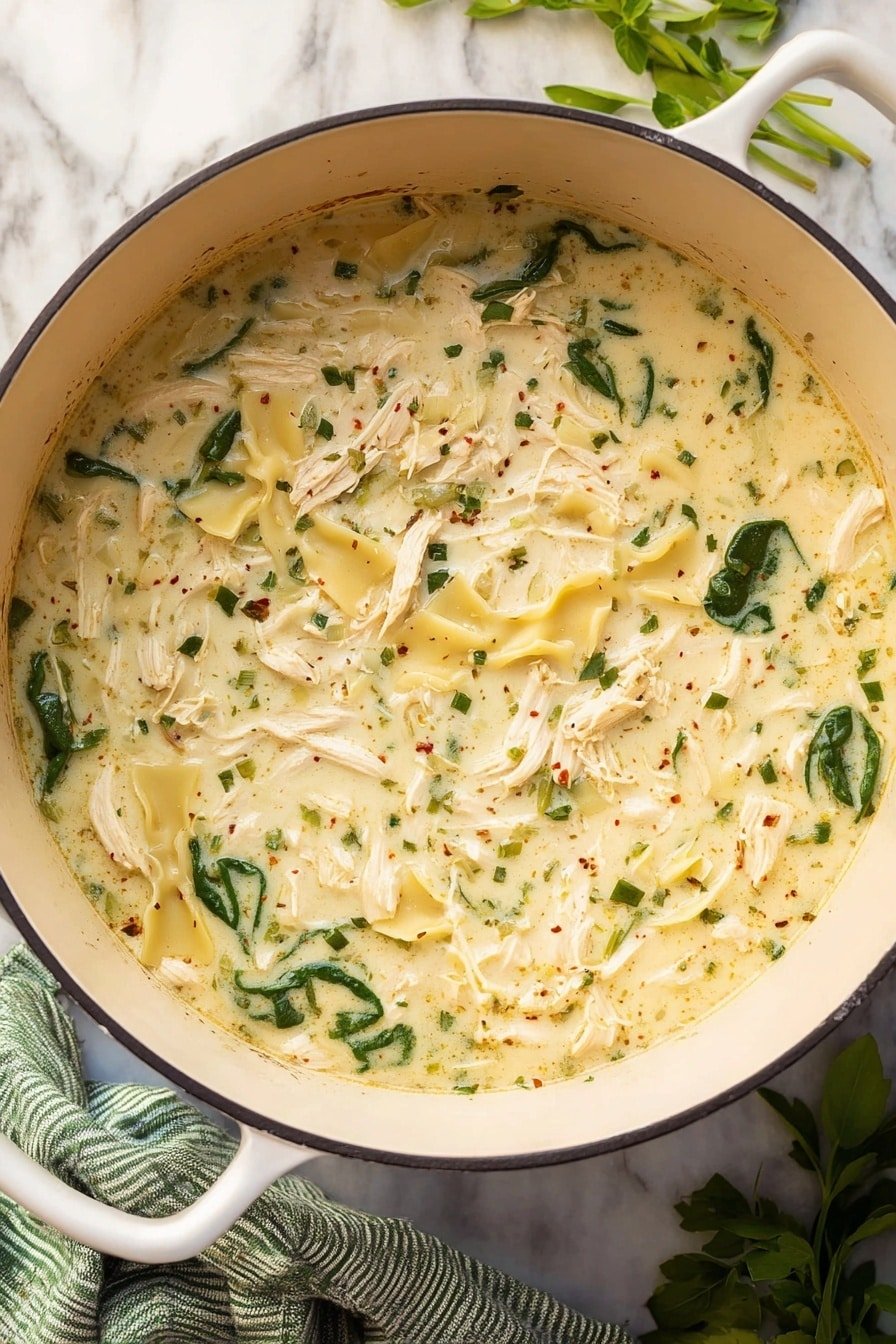 A large white pot filled with creamy soup containing three main layers: shredded white chicken pieces spread evenly through the soup, yellow wavy-edged pasta scattered throughout, and dark green spinach leaves adding color contrast. The soup base is thick and pale yellow with visible herbs and spices speckled across the surface. The pot sits on a white marbled surface, next to a green and white striped cloth, with some green leafy herbs placed nearby. Photo taken with an iphone --ar 2:3 --v 7