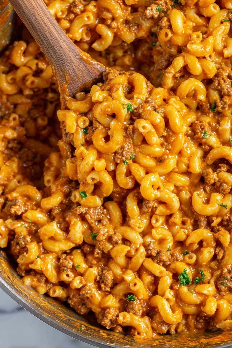 This image shows a close-up of a pan filled with cooked elbow macaroni mixed with ground meat in a thick orange sauce. The macaroni pieces are well coated with the sauce, and small chunks of cooked ground meat are spread evenly throughout. Some small green parsley pieces sprinkle across the dish adding a touch of color contrast. A wooden spoon is scooping a portion of the macaroni mixture, showing the texture and thickness of the sauce. The pan's inside edges show some residue of the sauce. The background is a white marbled texture. photo taken with an iphone --ar 2:3 --v 7