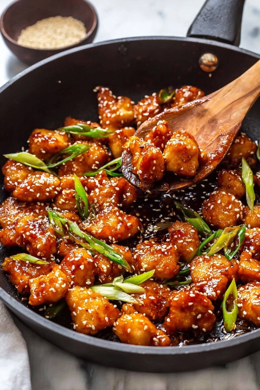 A black pan filled with many pieces of golden-brown cooked chicken chunks, each covered in a shiny, sticky sauce, scattered with white sesame seeds and bright green sliced scallions on top. A wooden spoon with a hole in the middle is lifting some chicken pieces, showing their glossy texture and orange-brown color. In the background, a small dark bowl holds a small pile of sesame seeds. The pan rests on a white marbled surface photo taken with an iphone --ar 2:3 --v 7