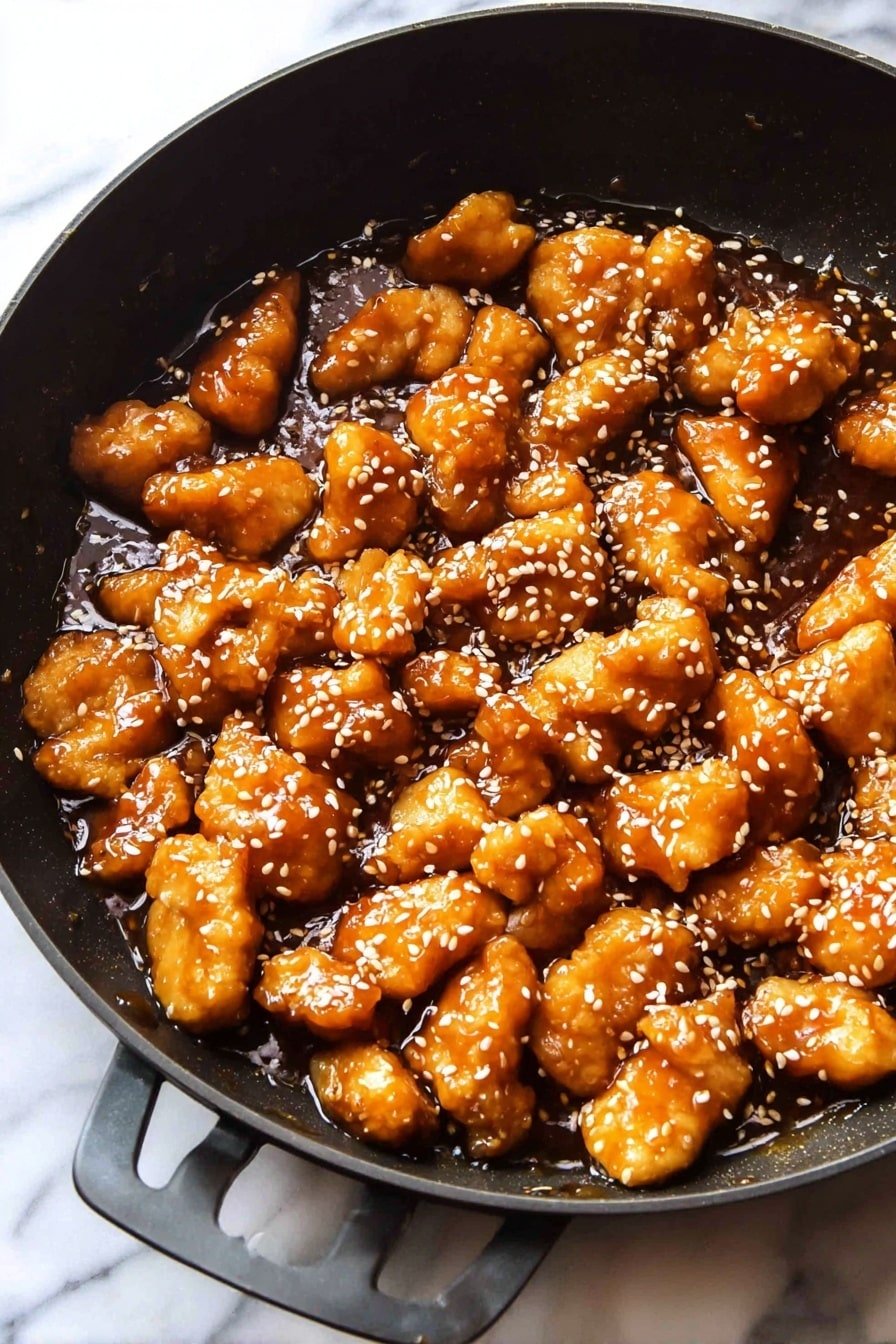 The image shows a black pan filled with small pieces of golden brown chicken coated in a shiny, thick sauce. The chicken pieces are scattered evenly and sprinkled with small white sesame seeds. The sauce looks sticky and rich, pooling slightly at the bottom of the pan. The pan rests on a white marbled surface. Photo taken with an iphone --ar 2:3 --v 7