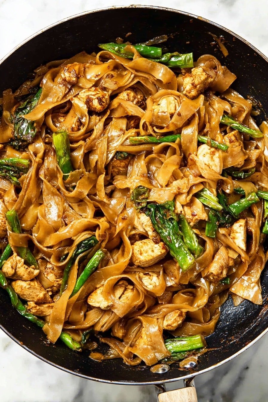 This dish shows a black pan filled with wide, flat rice noodles that have a light brown color from sauce coating them. Mixed throughout are pieces of cooked chicken, which are light golden brown and somewhat chunky. Green vegetables, including leafy greens and snap peas or green beans, add bright spots of color. The noodles and chicken are slightly glossy, indicating a sauce, and the dish has some dark caramelized bits. The pan sits on a white marbled surface, and a woman’s hand holds the pan’s handle. Photo taken with an iphone --ar 2:3 --v 7