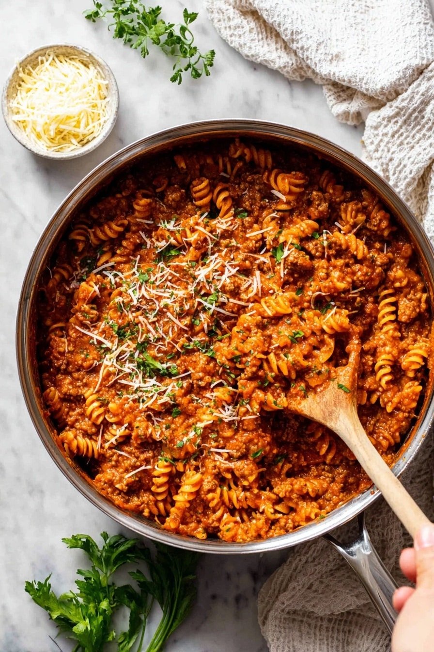 A large, silver pot filled with a thick, reddish-orange pasta sauce mixed with spiral pasta and ground meat. The sauce has a rich, slightly chunky texture and is sprinkled with small green herb pieces and thin shreds of white cheese on top. A woman's hand holds a large wooden spoon, lifting a scoop of the pasta mixture slightly above the pot's surface. Around the pot, there is a small white bowl with shredded cheese, a few green herb sprigs, and a white cloth, all placed on a white marbled surface. photo taken with an iphone --ar 2:3 --v 7