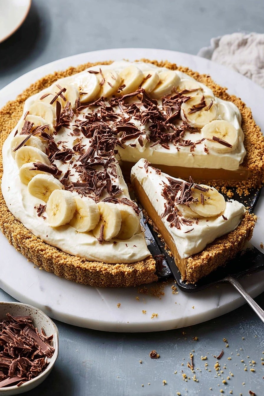 The image shows a round tart with four visible layers sitting on a white marble surface. The bottom layer is a golden brown crumbly crust forming a thick edge around the tart. Above that is a smooth, caramel-colored filling. Next, there is a layer of thin, evenly spaced banana slices that peek out from under a thick, creamy white topping. The top layer is decorated with curls and small shreds of dark chocolate scattered over the cream. One slice is being lifted from the tart with a black utensil, showing the distinct layers inside. Nearby, there is a white bowl with dark chocolate curls on the same white marble surface. Photo taken with an iphone --ar 2:3 --v 7