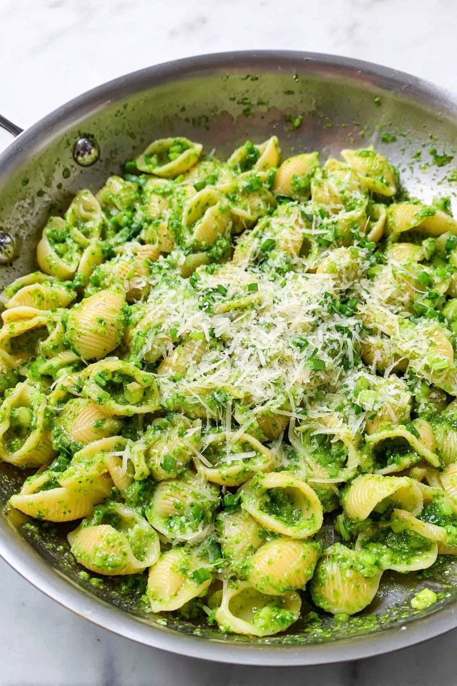 A silver pan filled with small shell pasta coated in a bright green pesto sauce, with bits of herbs and garlic visible in the sauce. On top of the pasta, there is a sprinkle of finely grated light yellow Parmesan cheese, adding a soft textured layer. The dish sits on a white marbled surface. photo taken with an iphone --ar 2:3 --v 7