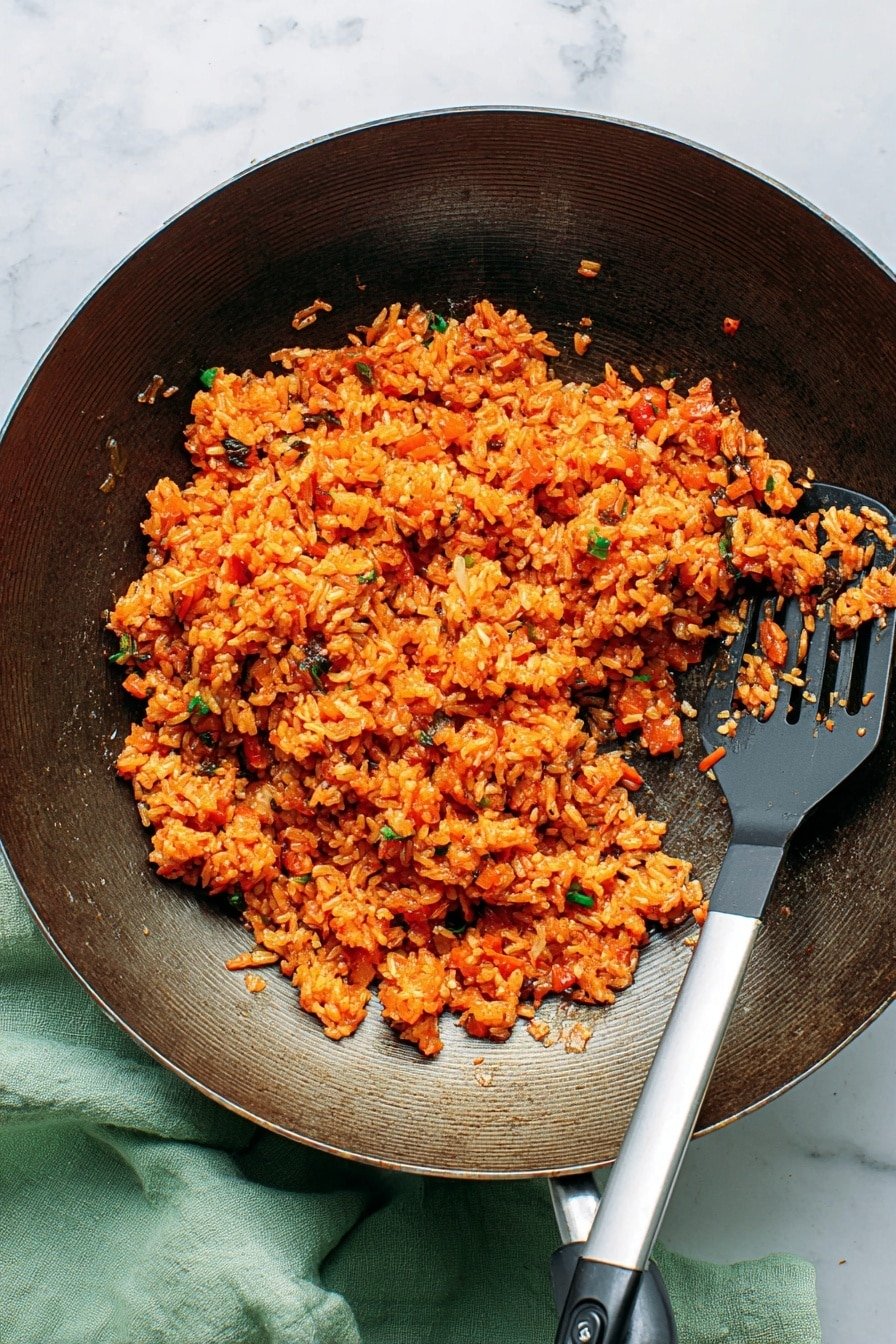 A dark metal wok filled with bright orange cooked rice mixed with small pieces of vegetables and herbs, showing a textured and slightly oily surface. The rice is piled toward the center-left side of the wok, with some bits scattered around, creating an uneven but full layer. A metal spatula with a black-and-white handle rests inside the wok on the right side, slightly under the rice. The wok sits on a white marbled surface, with a soft green cloth partially visible underneath on the left side. Photo taken with an iphone --ar 2:3 --v 7