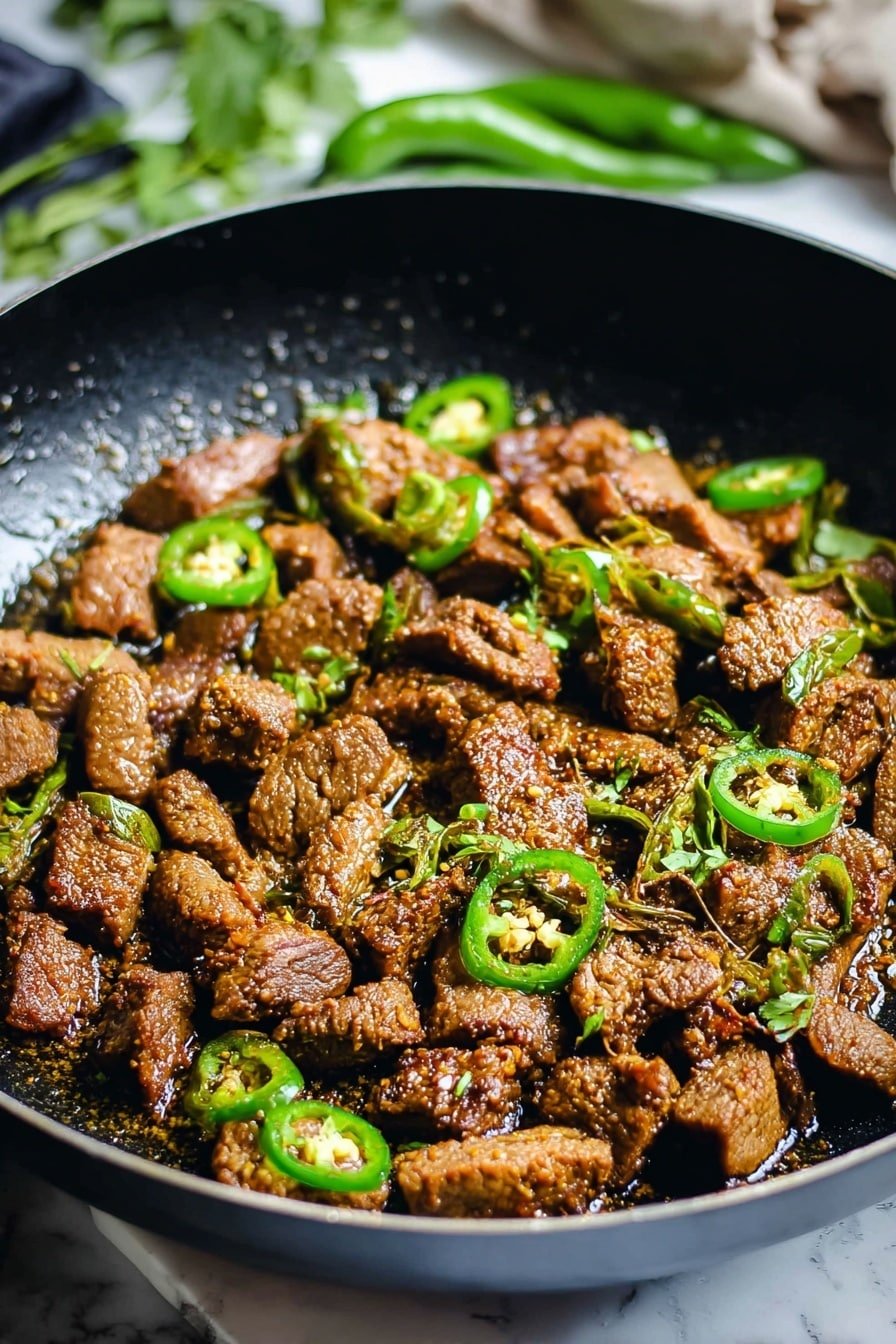 The image shows a black frying pan filled with small, brown, cooked meat pieces that have a slightly crispy texture. Scattered among the meat are thin, sliced green chili rings adding a fresh, bright green color contrast. The pan sits on a surface with a white marbled texture, and in the background, there are some green leaves which suggest fresh herbs. The meat glistens with a light coating of sauce or oil, making the dish look rich and savory. Photo taken with an iphone --ar 2:3 --v 7