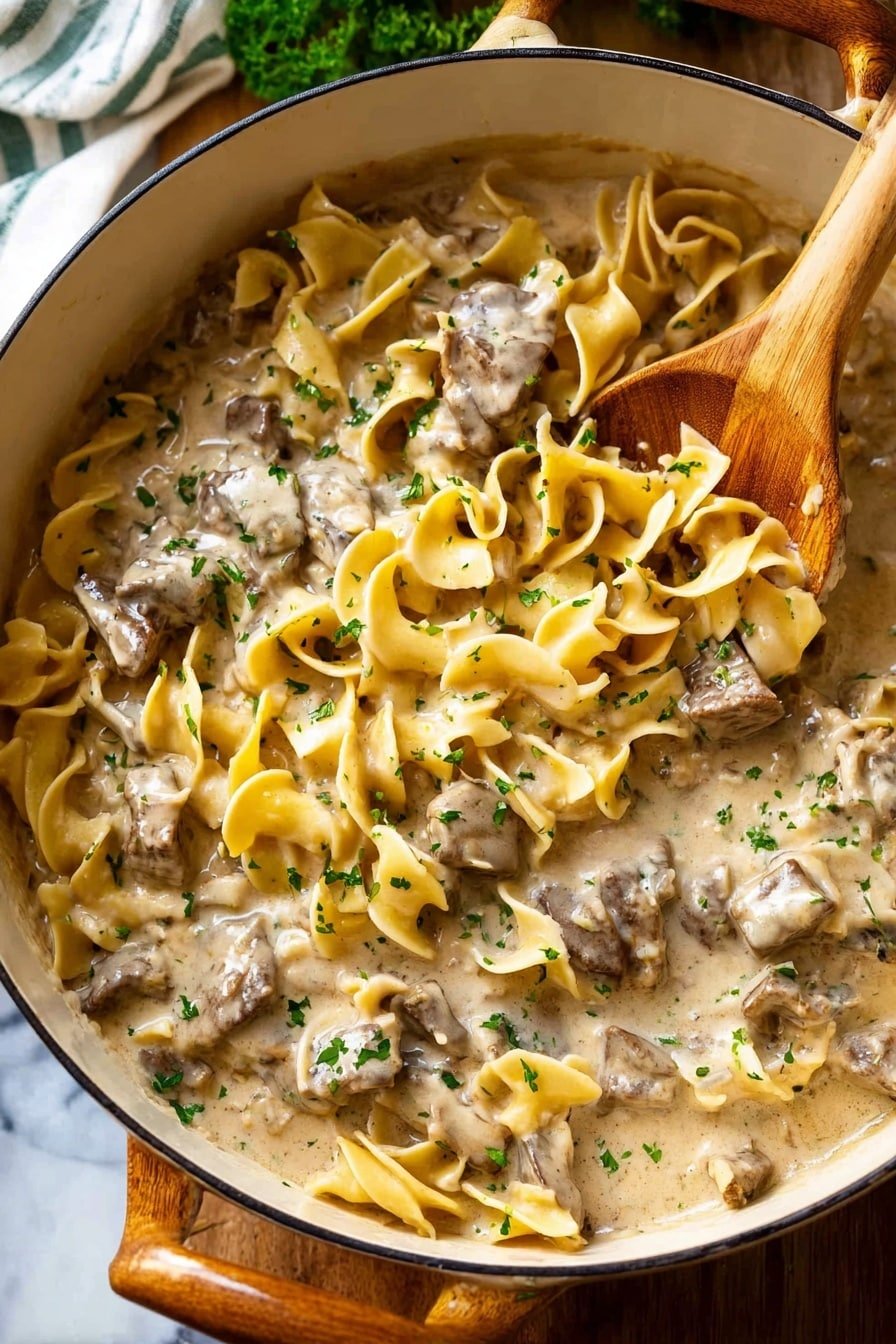 A close-up view of a creamy beef stroganoff dish in a white round pot with a wooden handle and a wooden spoon partially submerged. The dish has two main layers: wide, wavy egg noodles in pale yellow and tender chunks of beef mixed in a thick, light beige creamy sauce. Small sprinkled green herbs add a touch of color on top. The pot is placed on a wooden surface with a glimpse of a white marbled texture and some greens in the background. photo taken with an iphone --ar 2:3 --v 7