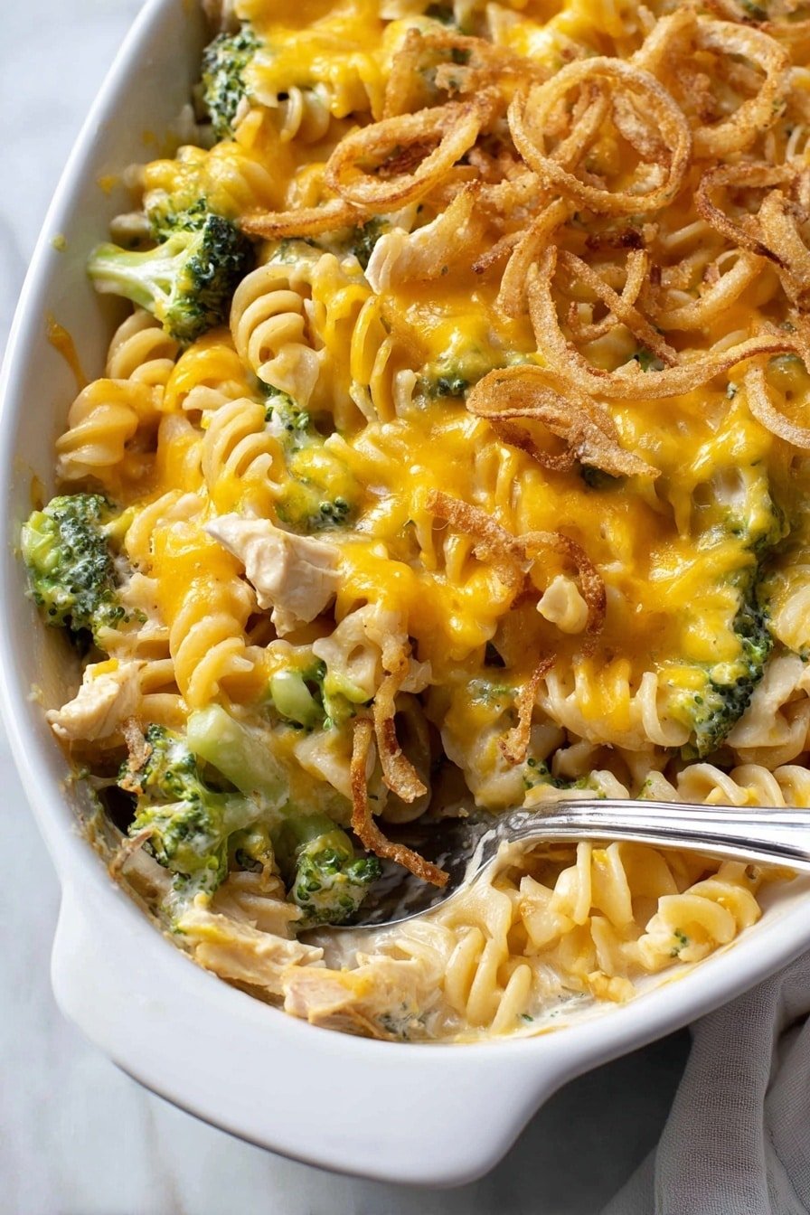 This is a close-up of a white dish filled with a layered pasta casserole. The first layer is creamy pasta spirals mixed with small green broccoli pieces. On top of that, there is a bright yellow melted cheese layer that covers the pasta and broccoli unevenly. Scattered on the surface are crispy light brown fried onion rings that add texture. A silver spoon is inside the dish, resting on the right side, partially scooping the pasta mix. The background shows a white marbled surface with a soft light setting. photo taken with an iphone --ar 2:3 --v 7