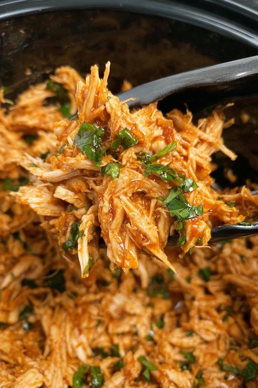 A close-up view of shredded orange-brown chicken mixed with small green herb leaves, held by silver tongs inside a black container, showing texture that is soft and fibrous with some moist sauce coating; the background and other parts of the container show more of the same shredded chicken. Photo taken with an iphone --ar 2:3 --v 7