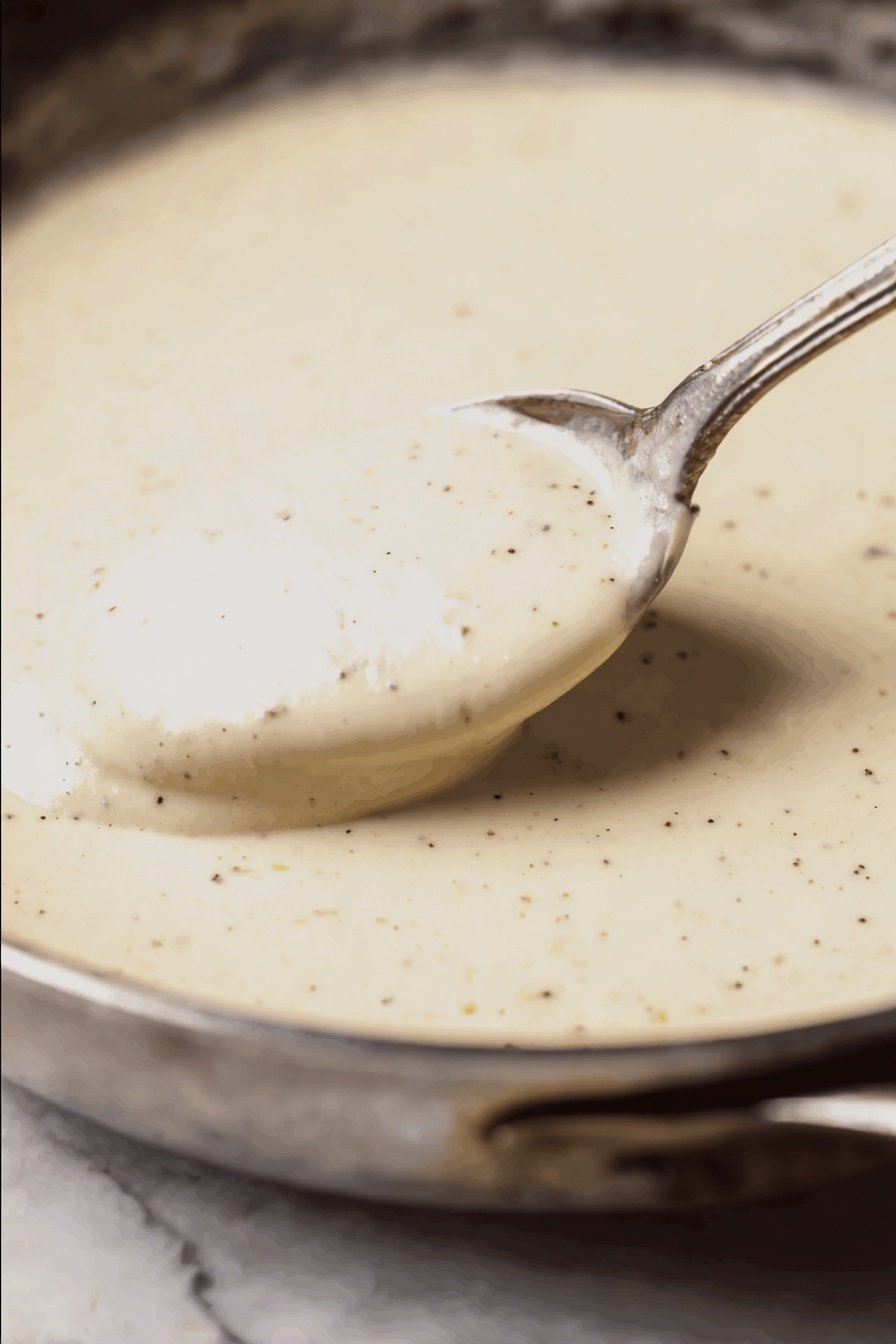 A close-up of a white plate with a thick slice of meatloaf cut in half showing two layers of brown, textured meat. A creamy white sauce with pepper specks covers the meatloaf and drips down the sides. A silver fork is stuck into the top layer, lifting it slightly. Behind the meatloaf, there is a mound of light-colored mashed potatoes with a soft texture. The plate is set on a white marbled surface. Photo taken with an iphone --ar 2:3 --v 7