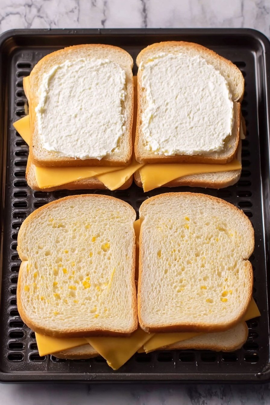 Two pieces of toasted white bread each topped with a layer of melted cheese that is light yellow with golden brown spots, showing a slightly crispy texture. The slices have a light golden crust and soft center beneath the cheese. Each slice is placed on a white plate with a thin dark rim, sitting on a white marbled surface. The focus is sharp on the slice in the foreground with the second slice slightly blurred in the background. Photo taken with an iphone --ar 2:3 --v 7