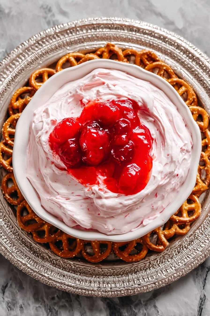 A white bowl sits in the middle of a round wooden tray filled with many small, twisted pretzels forming a detailed outer ring. Inside the bowl, there is one main layer of smooth, fluffy light pink cream with soft, swirled texture. On top, there is a bright red glossy layer of fruit sauce mixed with whole pieces of fruit, creating a shiny contrast to the cream below. The background shows a white marbled surface. photo taken with an iphone --ar 2:3 --v 7