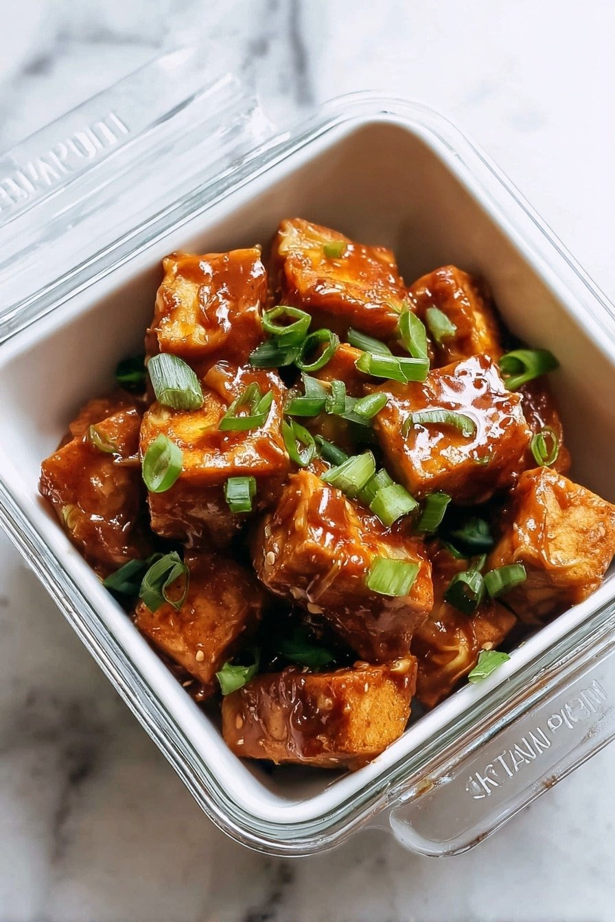 A close-up view of a square clear glass container filled with golden-brown crispy tofu cubes covered in a shiny, thick reddish-brown peanut sauce, garnished with bright green sliced scallions scattered on top. The container rests on a white marbled surface with a partially open clear glass lid showing a label that reads