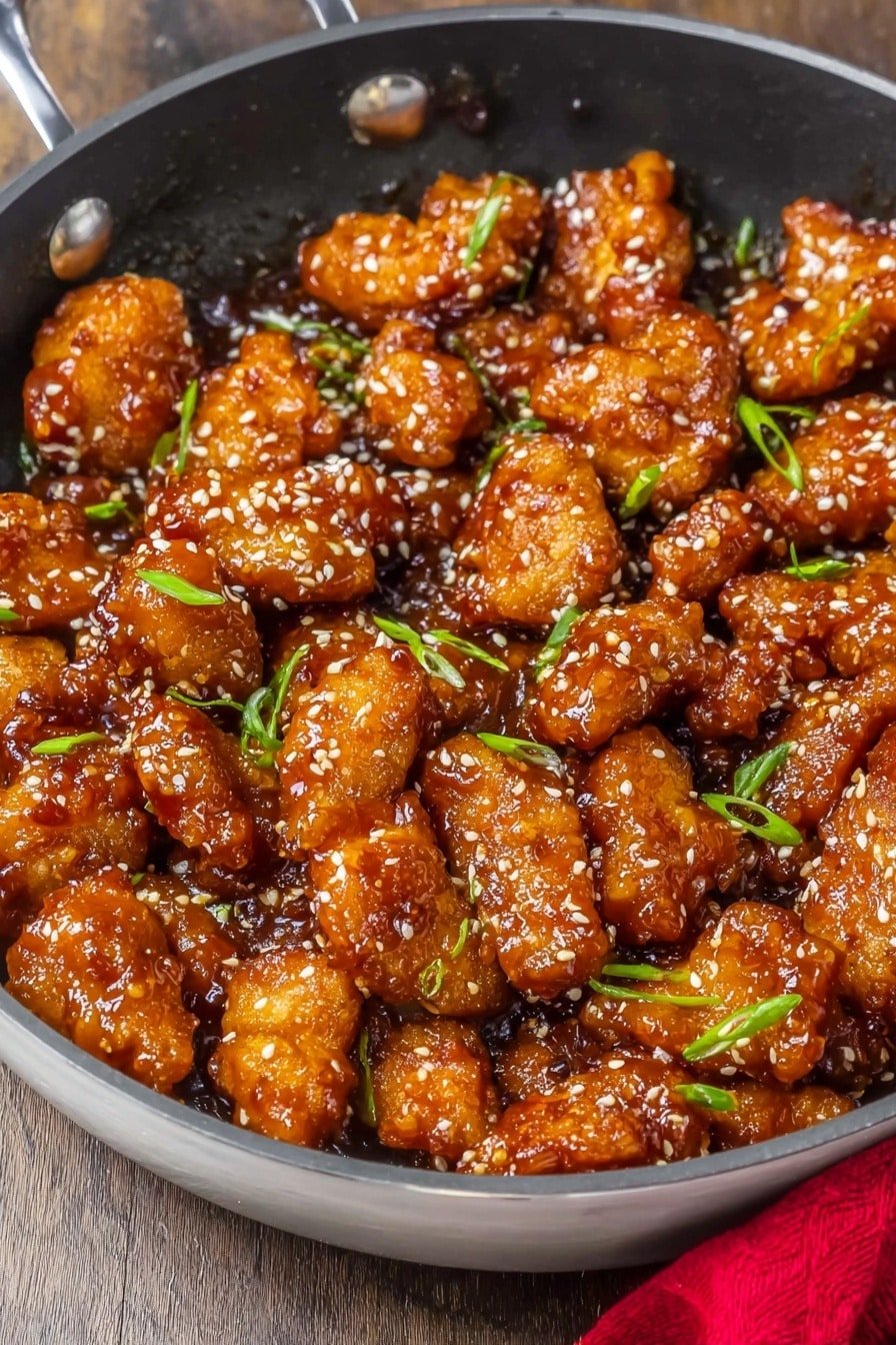 A close-up view of a black cast iron skillet filled with many pieces of glazed orange-brown chicken wings, each piece covered in a shiny, sticky sauce and sprinkled with light sesame seeds and small green onion slices, creating a mix of textures on the surface. The skillet sits on a white marbled surface and part of a white cloth napkin is visible near the top right corner. The chicken pieces vary in size and are densely packed, showing their crispy edges and soft inside. photo taken with an iphone --ar 2:3 --v 7