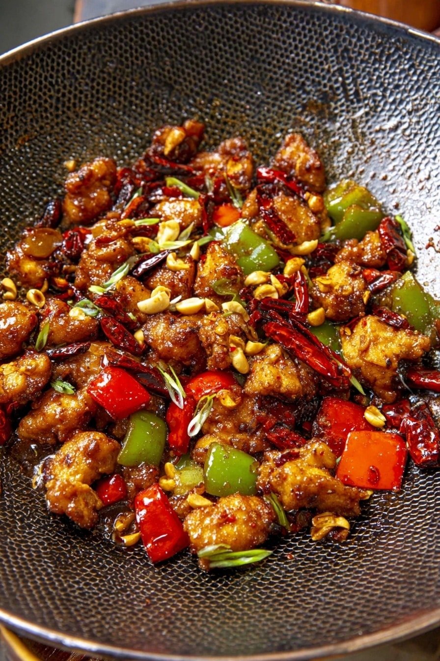 The dish shows many pieces of cooked meat with a brown, crispy texture mixed with bright green and red bell pepper chunks, all in a dark pan with a honeycomb pattern. Scattered on top are whole dried red chili peppers, small pieces of chopped peanuts, and tiny green onion slices, adding detail and color contrast. The meat and vegetables look shiny with sauce that makes them look juicy and flavorful. The close-up view focuses on the stir-fry inside the pan, with a wood texture barely visible around the edges. photo taken with an iphone --ar 2:3 --v 7