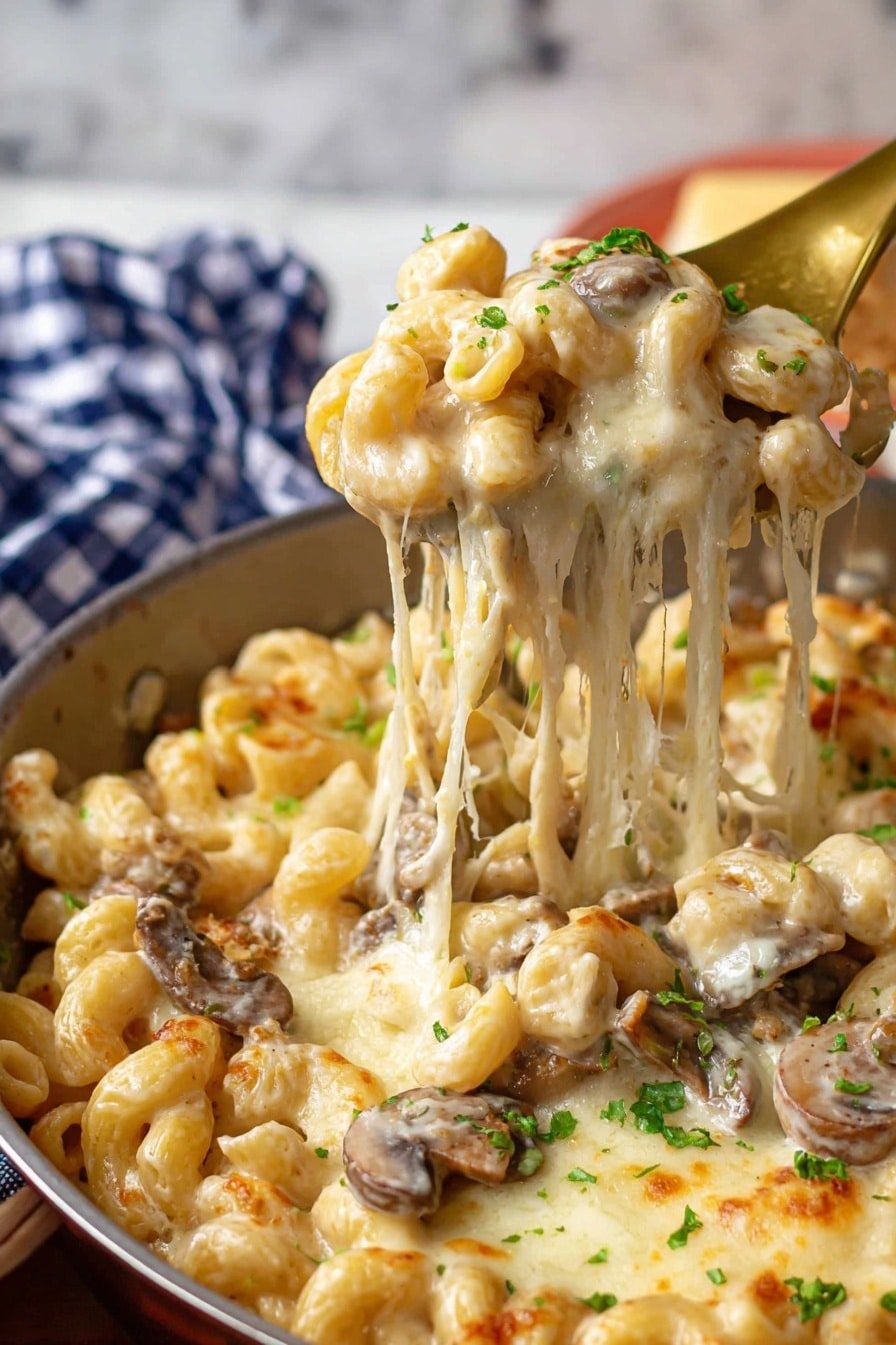 The image shows a close-up of a white pan filled with creamy macaroni pasta mixed with pieces of brown mushrooms, layered with melted, golden-brown cheese that stretches as a spoon lifts a portion from the pan. The pasta is covered in a smooth, creamy sauce with parsley sprinkled lightly on top for color. The background includes a white marbled surface with blurred red and white checkered fabric and green bell pepper. photo taken with an iphone --ar 2:3 --v 7