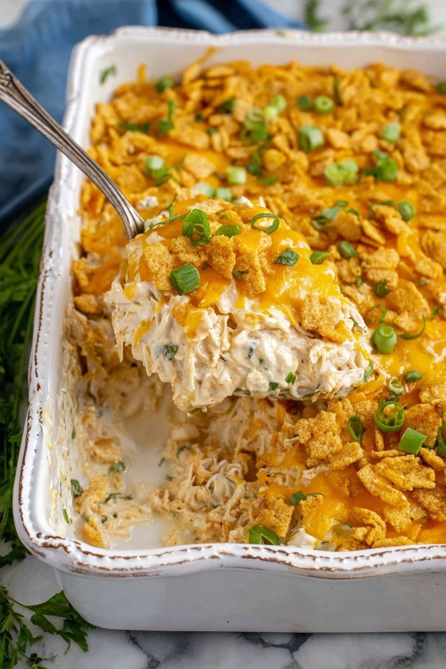 The dish is a creamy casserole in an ornate white rectangular baking dish with three visible layers. The bottom layer is shredded white meat chicken mixed with a creamy white sauce, visible where a golden spoon lifts a portion. The middle layer includes crunchy golden cracker pieces mixed throughout. The top layer is melted golden cheddar cheese sprinkled with sliced green onions and small green herb pieces. The casserole sits on a white marbled surface with some green herbs and a blue cloth in the background. The photo taken with an iphone --ar 2:3 --v 7