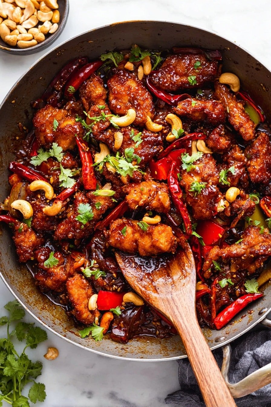 A large pan filled with a colorful stir-fry dish featuring several pieces of golden brown, crispy fried chicken coated in a thick, shiny dark brown sauce. Mixed in are bright red slices of bell pepper and whole dried red chili peppers scattered evenly around the chicken. Small crescent-shaped light tan cashews add texture and contrast, while tiny green cilantro leaves are sprinkled across the top for a fresh touch. A wooden spoon resting inside the pan scoops up some chicken, peppers, cashews, and sauce. The pan sits on a white marbled surface, with a few fresh cilantro sprigs nearby and a small bowl of crushed cashews visible in the background. Photo taken with an iphone --ar 2:3 --v 7