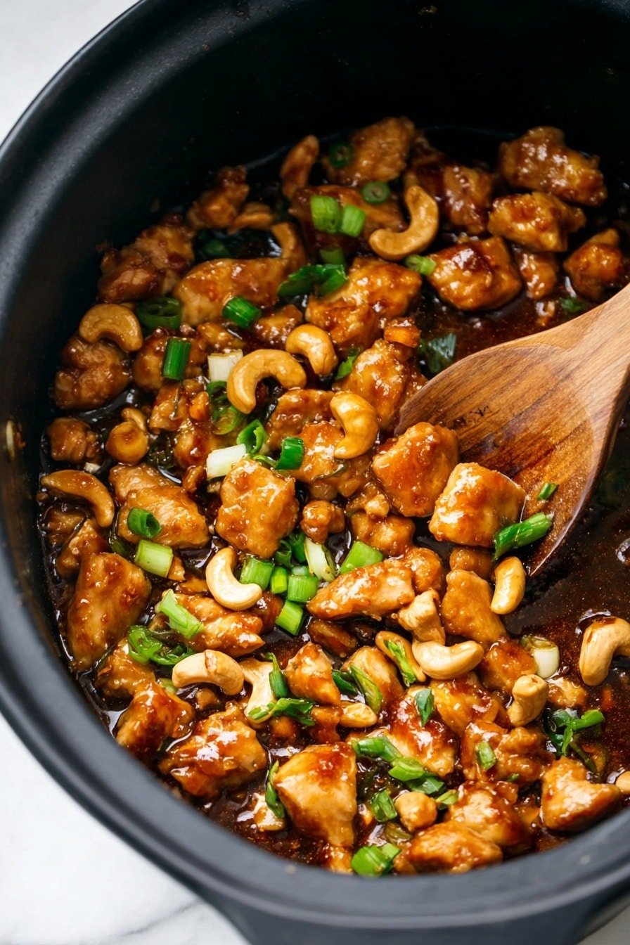 This image shows a close-up of a black pot filled with small, golden-brown pieces of cooked chicken covered in a shiny, thick brown sauce. Mixed in are light beige cashew nuts and small bright green sliced scallions scattered on top. A wooden spoon is partially buried in the food on the right side, lifting some pieces. The black pot sits on a white marbled surface. photo taken with an iphone --ar 2:3 --v 7
