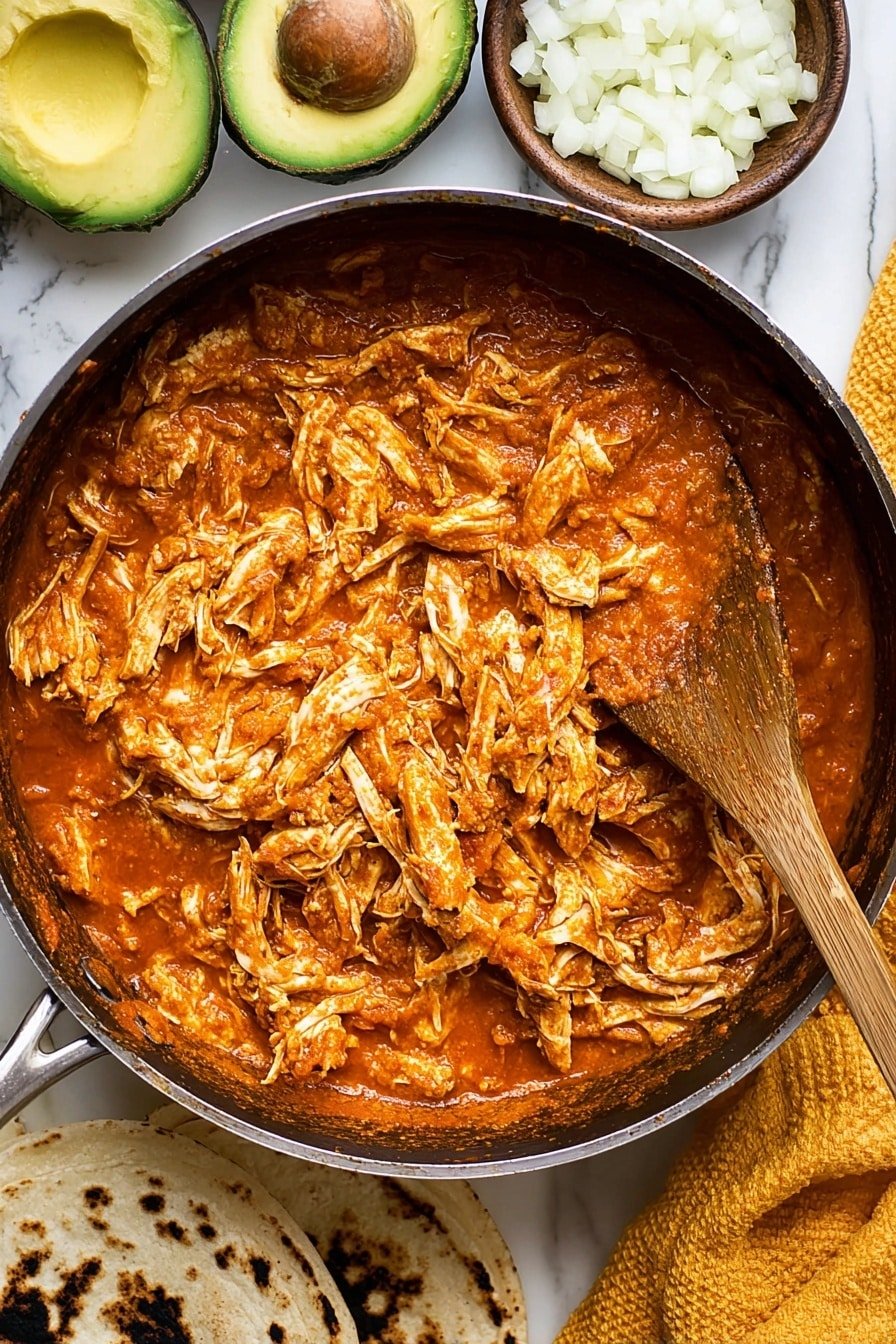 A black pan filled with shredded chicken mixed in a rich, orange-red sauce, giving it a moist and tender texture with some visible chicken fibers. A wooden spoon rests inside the pan, partially covered with sauce. Surrounding the pan on a white marbled surface are a few chopped white onions in a small bowl, a sliced avocado with a large pit showcasing its green and brown colors, a yellow cloth, and two warm, grilled tortillas with dark char marks. Photo taken with an iphone --ar 2:3 --v 7