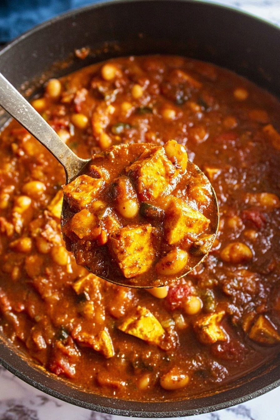 A close-up view of a thick stew in a black pot, showing a rich reddish-brown sauce filled with chunks of yellow tofu, white beans, diced tomatoes, and small pieces of dark green vegetables scattered throughout. A silver ladle lifts a portion of the stew, holding a mix of tofu, beans, and sauce with a shiny, smooth texture. The background is a white marbled texture with a hint of blue at the edges. photo taken with an iphone --ar 2:3 --v 7