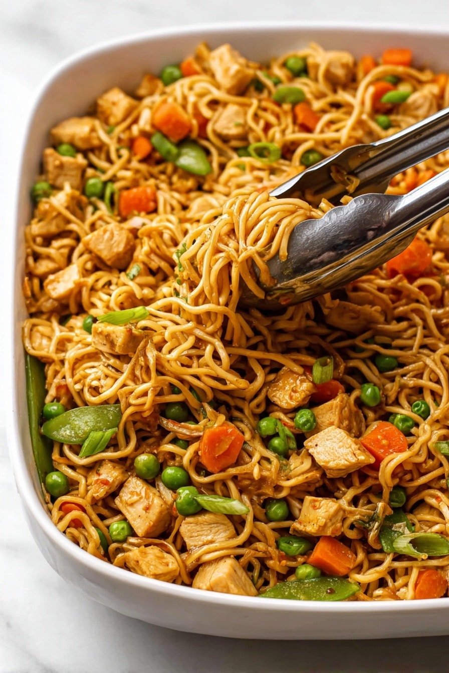 The image shows a white dish filled with cooked Asian-style noodles mixed with small cubes of cooked chicken, bright orange carrot pieces, green peas, and whole snap peas. The noodles are light brown, wispy, and slightly shiny from sauce, densely packed across the dish with some raised in a cluster being held by silver tongs. The green peas and carrots are scattered evenly, adding contrast to the noodle base, and small pieces of chopped scallions are visible on top. The dish sits on a white marbled surface, and the food looks warm and freshly served. photo taken with an iphone --ar 2:3 --v 7