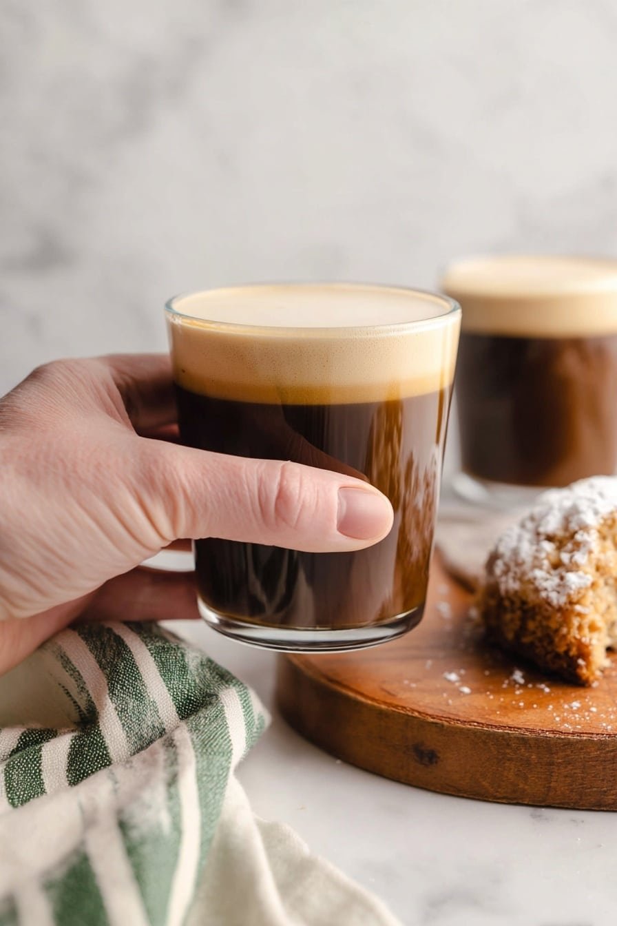 Two clear glasses filled about three-quarters with dark brown coffee topped with a thick layer of light tan cream. A woman's hand holds a spoon lifting the creamy topping, showing its soft and foamy texture over the first glass. Both glasses sit on a wooden board next to a square piece of pale yellow cake dusted with white powdered sugar. A clear bowl with a whisk and some cream is blurred in the background, all placed on a white marbled surface. A white and green cloth is partially visible at the bottom left corner. Photo taken with an iphone --ar 2:3 --v 7