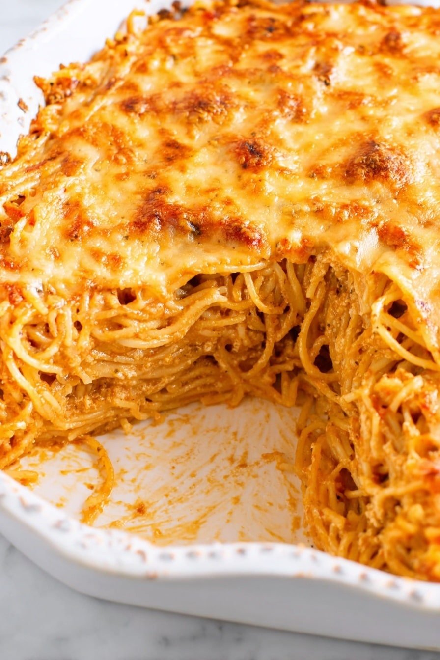 The image shows a baked spaghetti dish in an ornate white dish. The top layer is covered with melted golden cheese that looks slightly browned and bubbly. Below the cheese, there are multiple layers of cooked spaghetti noodles mixed with a light tomato sauce, visible as a reddish tint between the noodle layers. A section has been removed, revealing the spaghetti strands tightly packed and layered, showing a mix of light orange from the sauce and cream from the noodles. The dish is placed on a white marbled surface. photo taken with an iphone --ar 2:3 --v 7