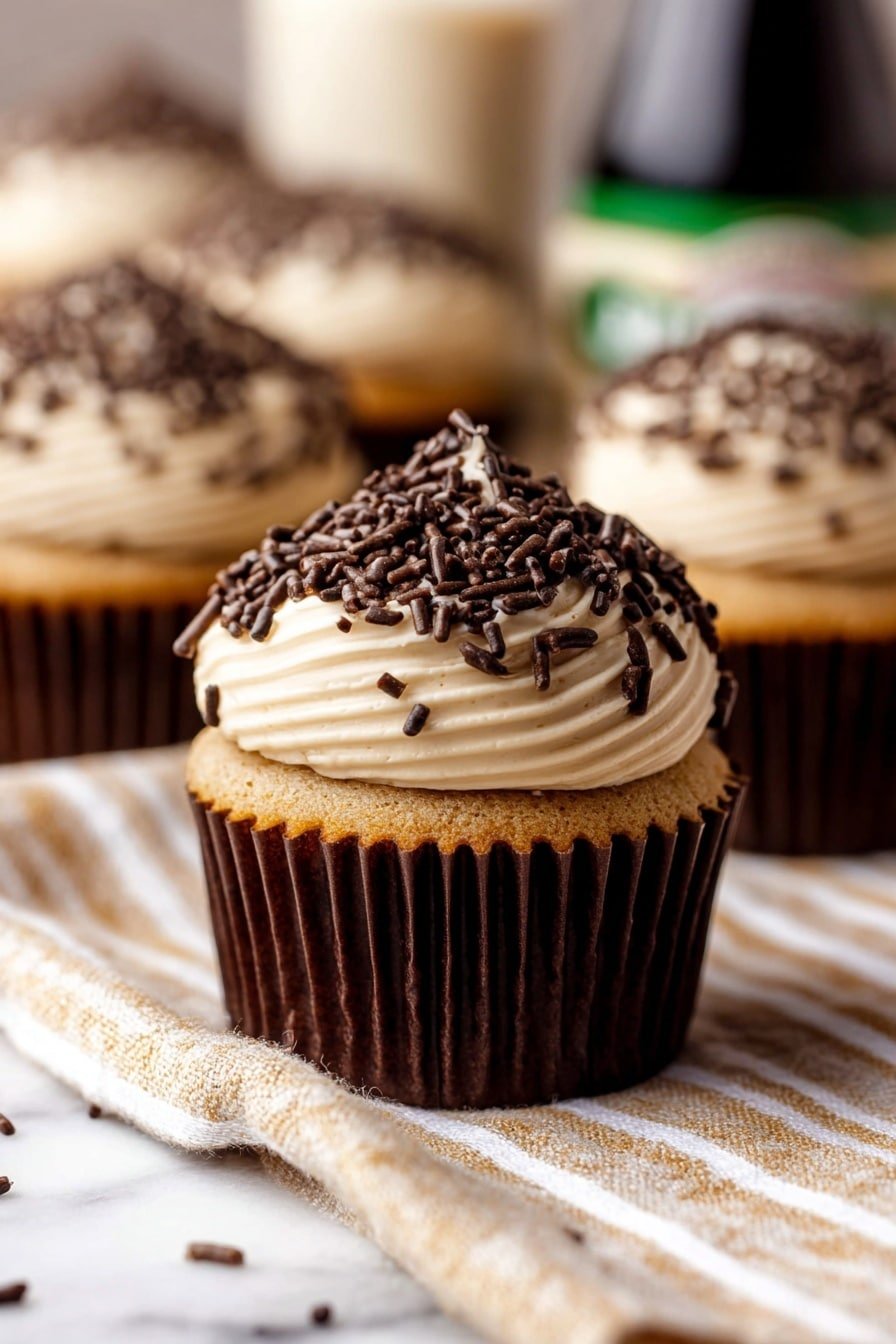 A close-up view of a light brown cupcake in a dark brown paper liner, topped with one thick layer of smooth, creamy light beige frosting that swirls into a rounded peak, generously covered with dark brown chocolate sprinkles spread all over the frosting. The cupcakes rest on a light tan and white striped fabric with a blurry background showing more similar cupcakes and an out-of-focus bottle, all placed on a white marbled texture. photo taken with an iphone --ar 2:3 --v 7