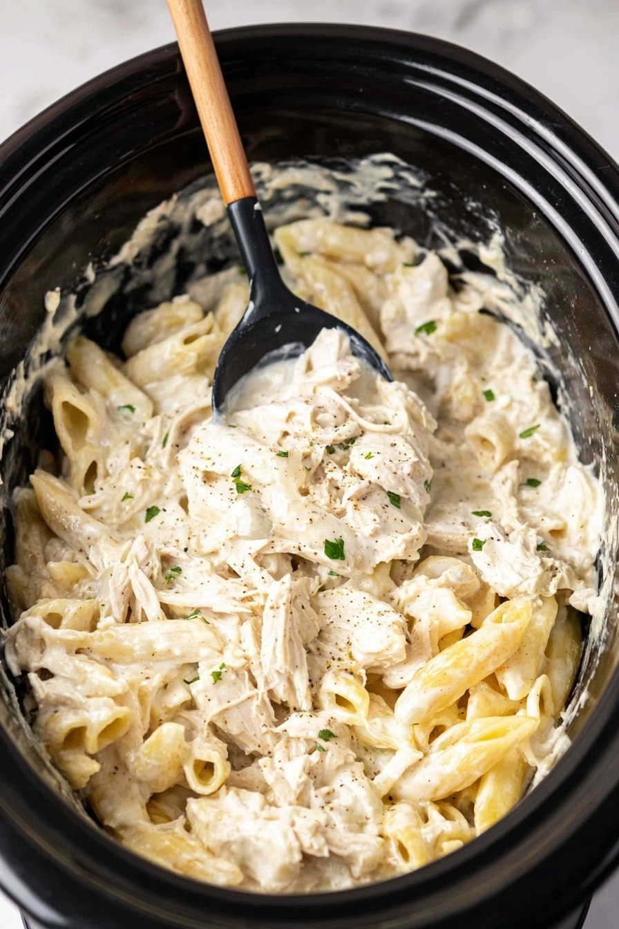 A close-up view of creamy pasta inside a black slow cooker, showing about two layers of thick white sauce-covered pasta mixed with shredded light beige chicken pieces, small bits of chopped green herbs, and black pepper specks. A wooden spoon is lifted in the center, stirring the pasta, with some sauce clinging to it. The pasta shapes include short, twisted tubes and flattened pieces with wavy edges, all coated in smooth, creamy white sauce. The slow cooker bowl shows some sauce stains around the inside edge. The setting is on a white marbled texture surface. Photo taken with an iphone --ar 2:3 --v 7