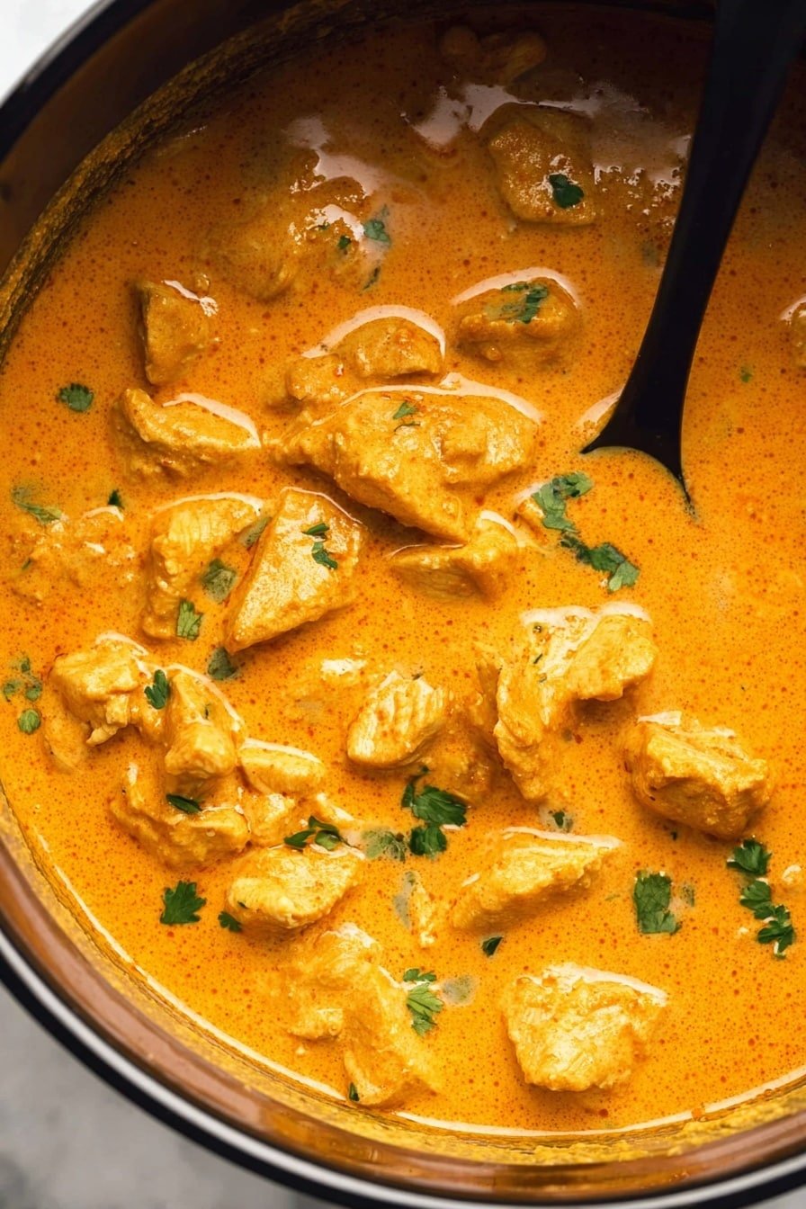 This is a close-up of a pot filled with thick, orange creamy curry sauce. Inside the sauce, there are many chunks of tender meat evenly spread throughout. The sauce has a smooth texture with small green herb pieces sprinkled on top. A black spoon is partially dipped into the curry on the right side of the pot. The pot is on a white marbled texture. photo taken with an iphone --ar 2:3 --v 7