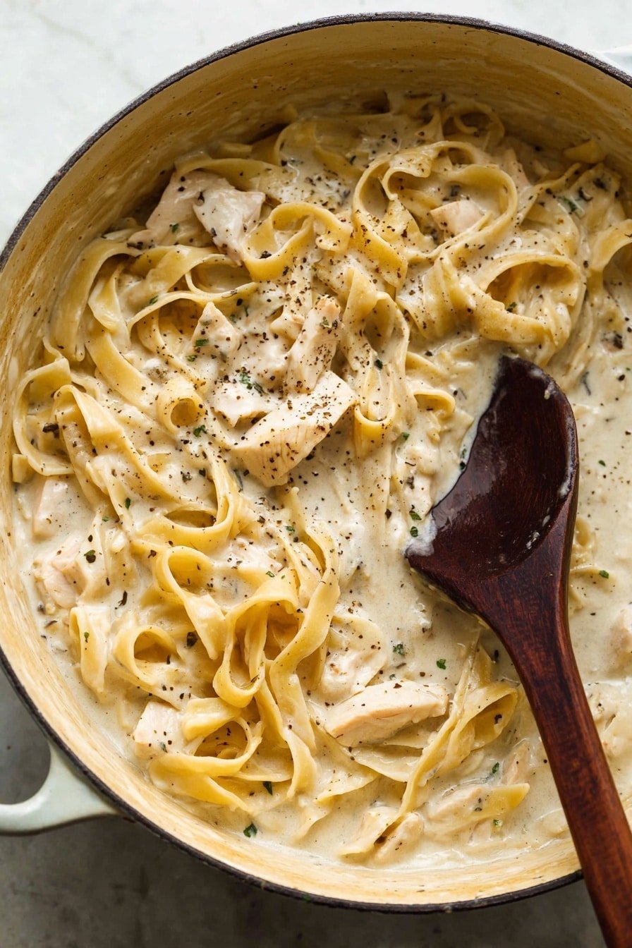 The image shows a close-up of a large cooking pot filled with creamy fettuccine pasta in a thick, pale beige sauce. The pasta noodles are wide and flat, layered unevenly throughout the pot, coated well with the smooth sauce dotted with black pepper and small green herbs. Small chunks of light, cooked chicken are mixed in among the pasta. A dark wooden spoon is partially submerged stirring the dish on the right side, with some sauce dripping off it. The inside of the pot is light beige with tiny scattered specks, and the whole scene is set on a white marbled texture. photo taken with an iphone --ar 2:3 --v 7