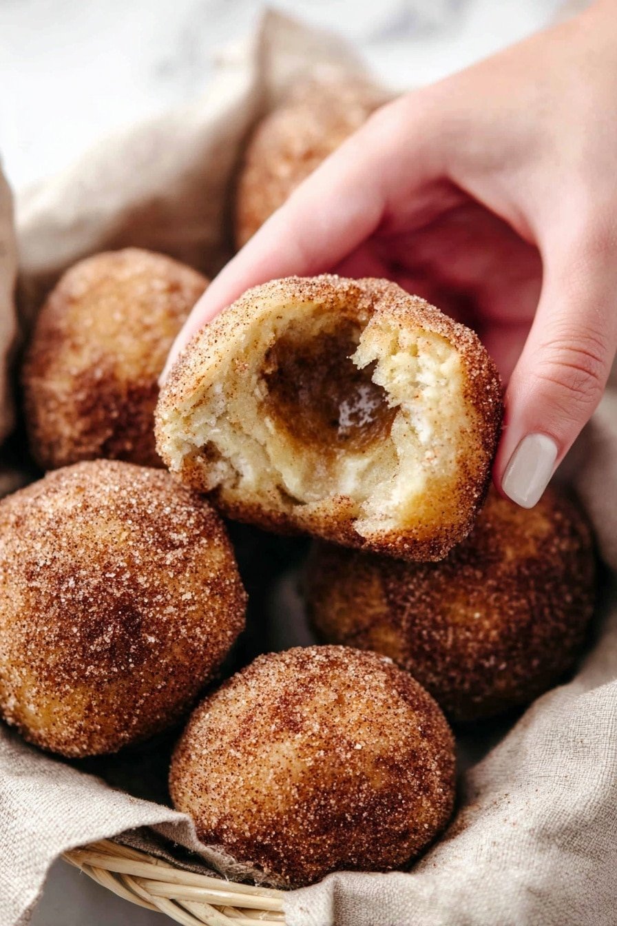 A basket lined with soft gray cloth holds a stack of round dough balls covered in a powdery brown cinnamon coating. Some dough balls are cracked open, showing inside layers of soft, light beige dough wrapped around a sticky, dark caramel-colored filling. The basket sits on a white marbled surface, with a few cinnamon sticks nearby, adding a warm, rustic touch to the scene. Photo taken with an iphone --ar 2:3 --v 7